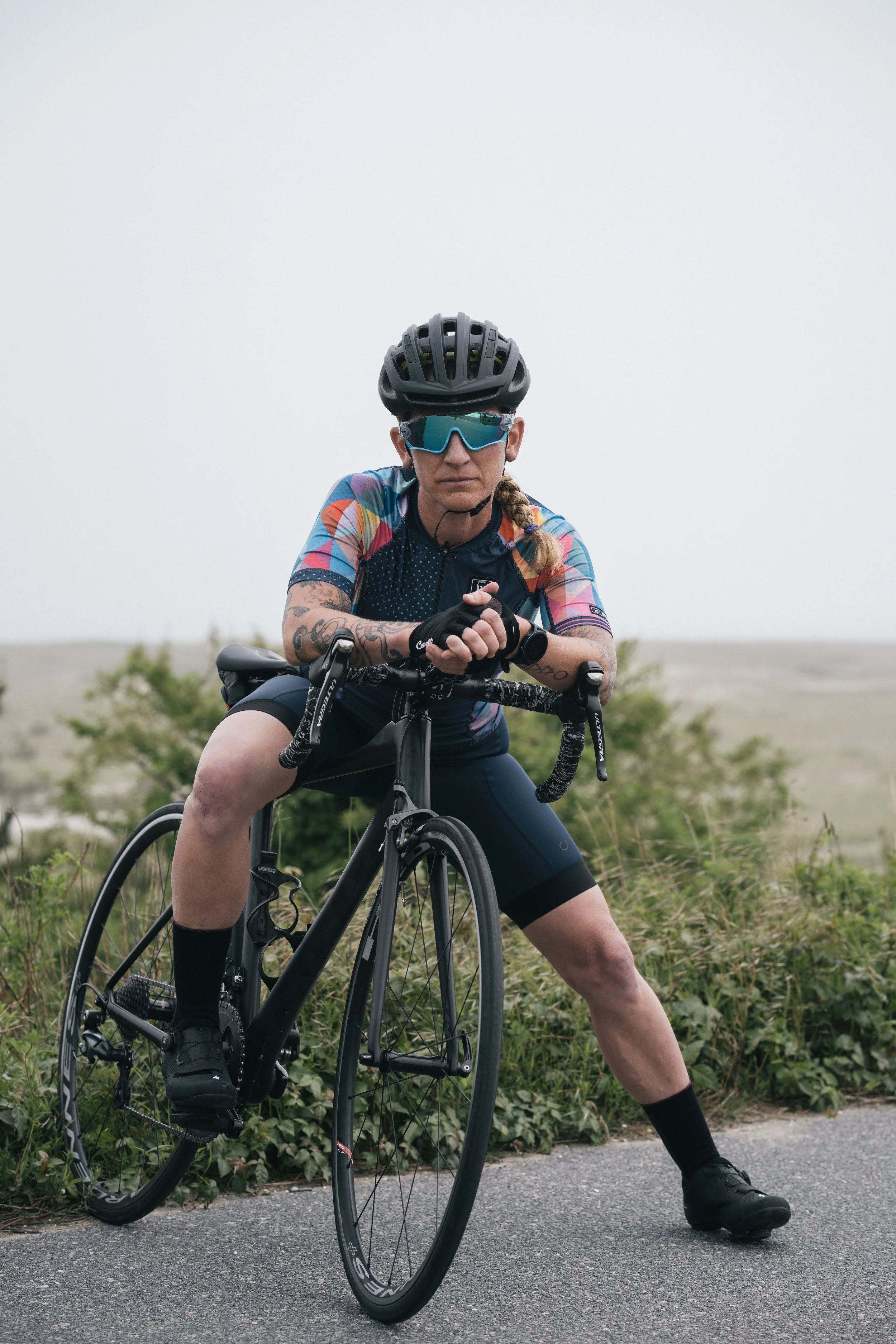 A person dressed in cycling gear, including a black helmet, blue-tinted sunglasses, and colorful cycling jersey, is sitting on a black road bicycle outdoors on a paved path surrounded by greenery, with an overcast sky in the background.