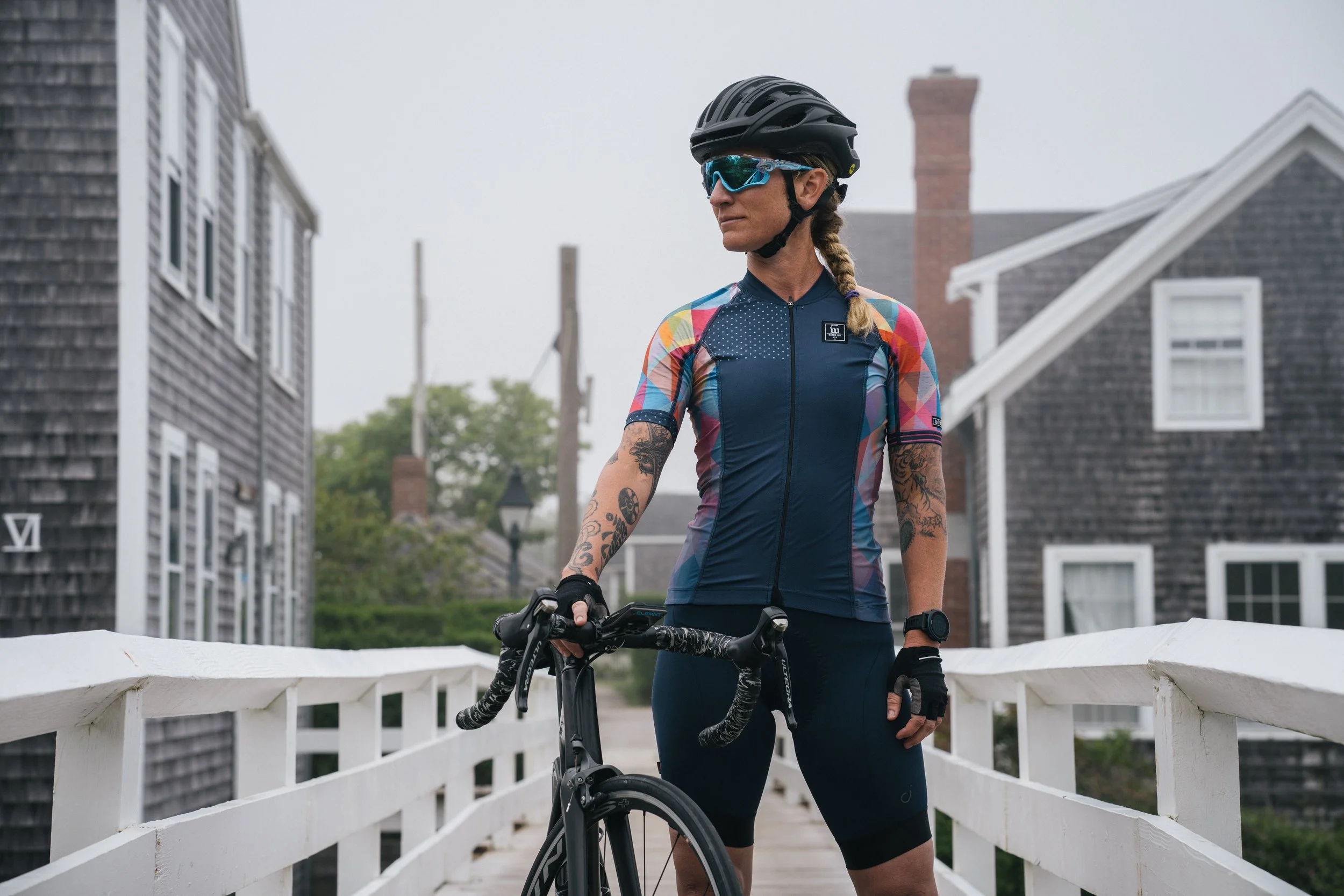 A woman wearing a black helmet, blue sunglasses, and a colorful cycling jersey stands beside her black road bike on a white bridge with wooden houses in the background, overcast sky.