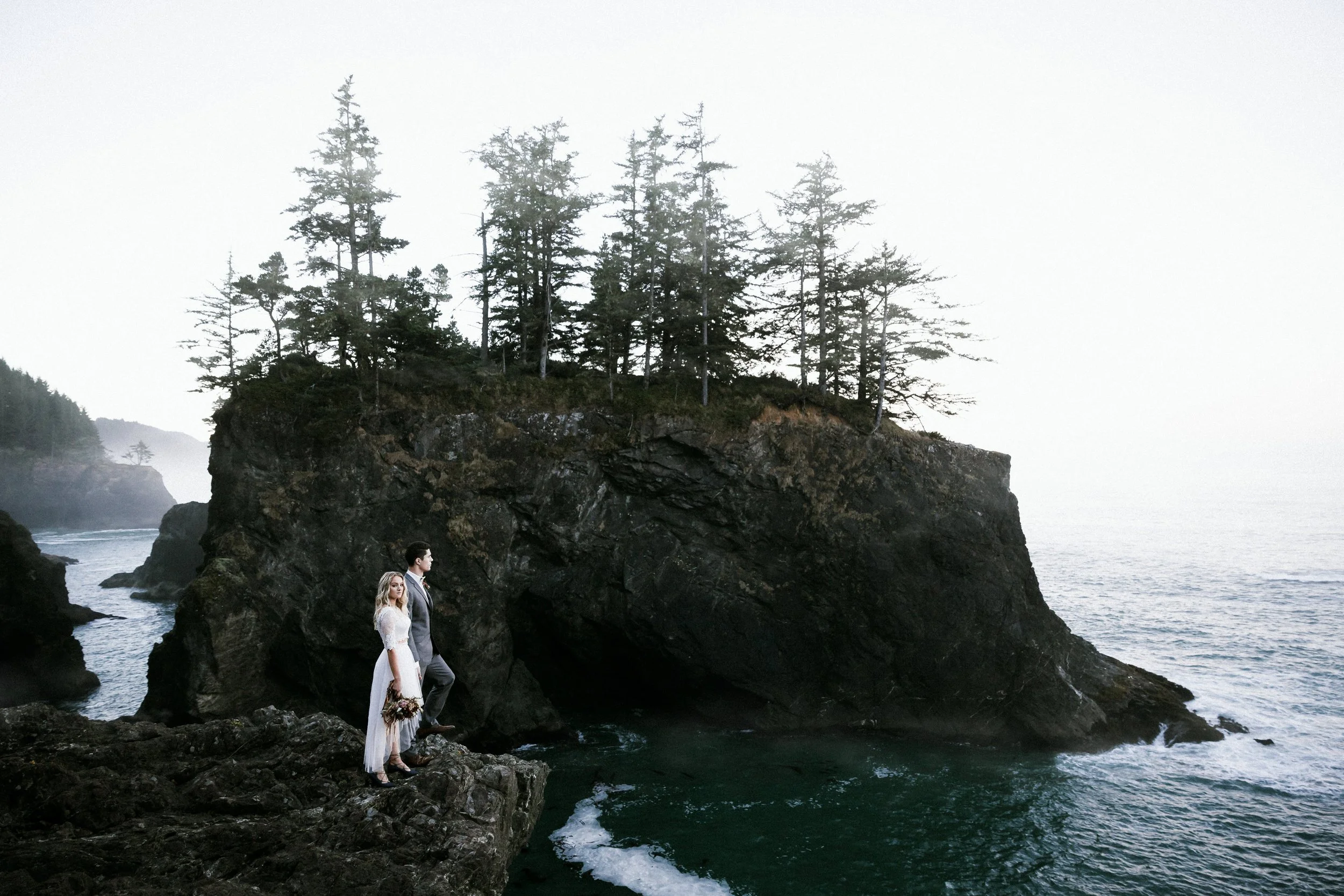 Bride standing on a dramatic rock arch over the ocean near rugged sea stacks, captured by Bend, Oregon wedding photographer Tony Gambino.