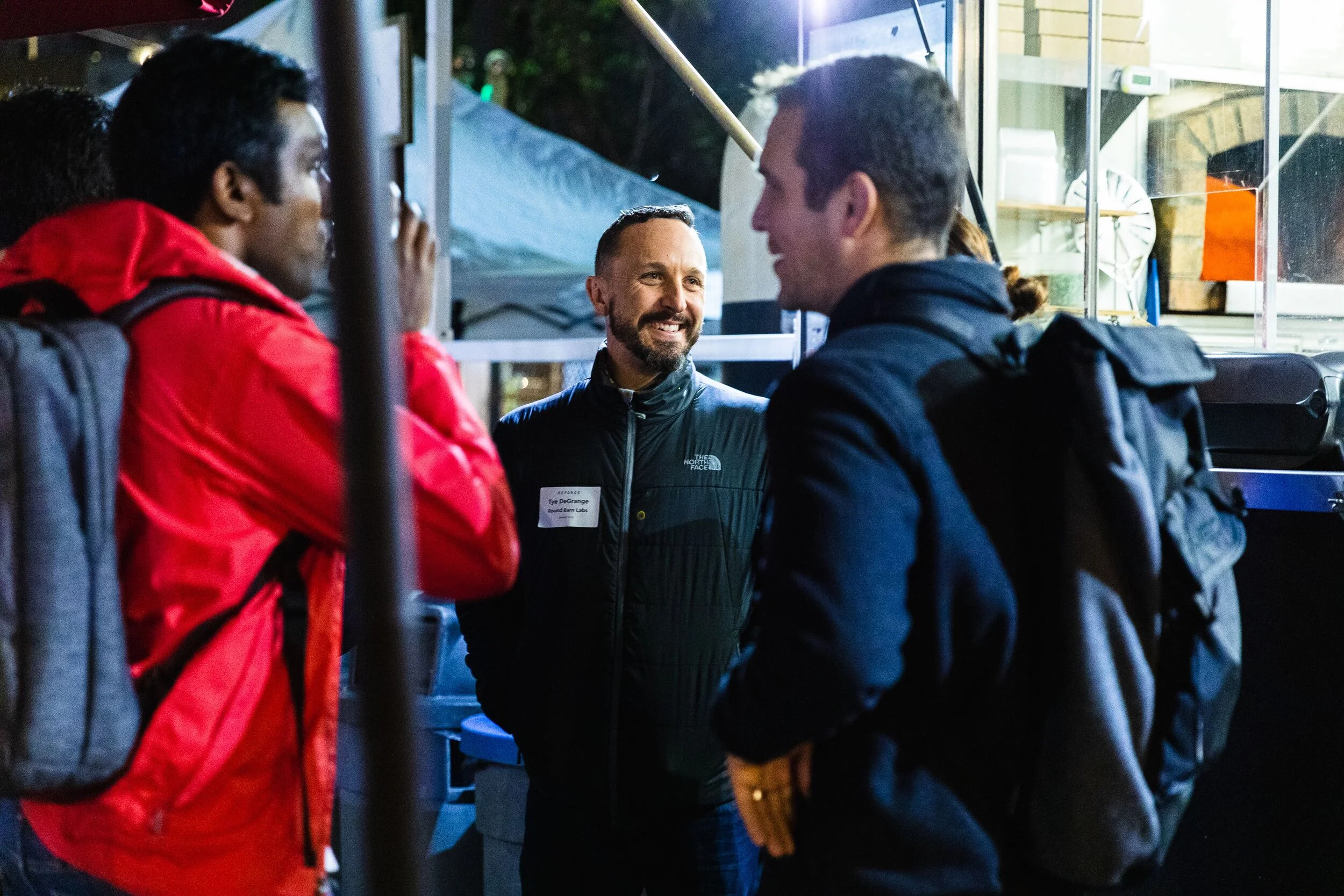 Three men conversing at an outdoor event, wearing backpacks and jackets, one has a name badge, near a stall or booth.