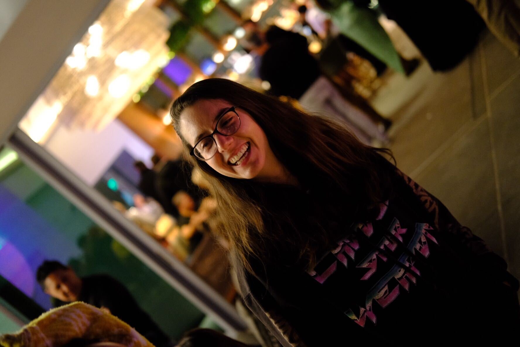 Woman smiling in a busy outdoor setting, wearing glasses and a shirt with neon text, with blurred people and lights in the background.