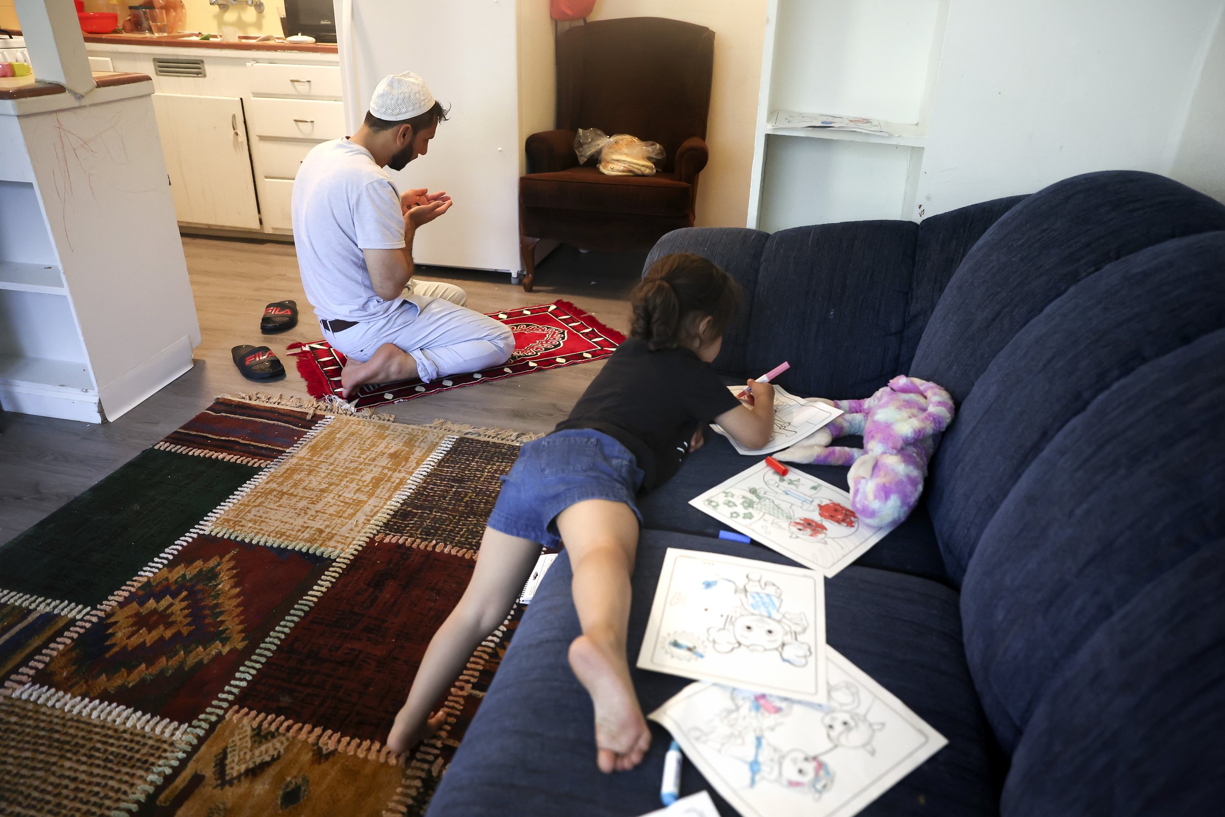  Zahra Mohammadi lays on the couch and colors while her father Najibullah Mohammadi prays in the kitchen  on Tuesday, May 3, 2022. Najibullah Mohammadi, his wife Susan, their two young children Zahra and Yasar, and their soon-to-be born third child Y