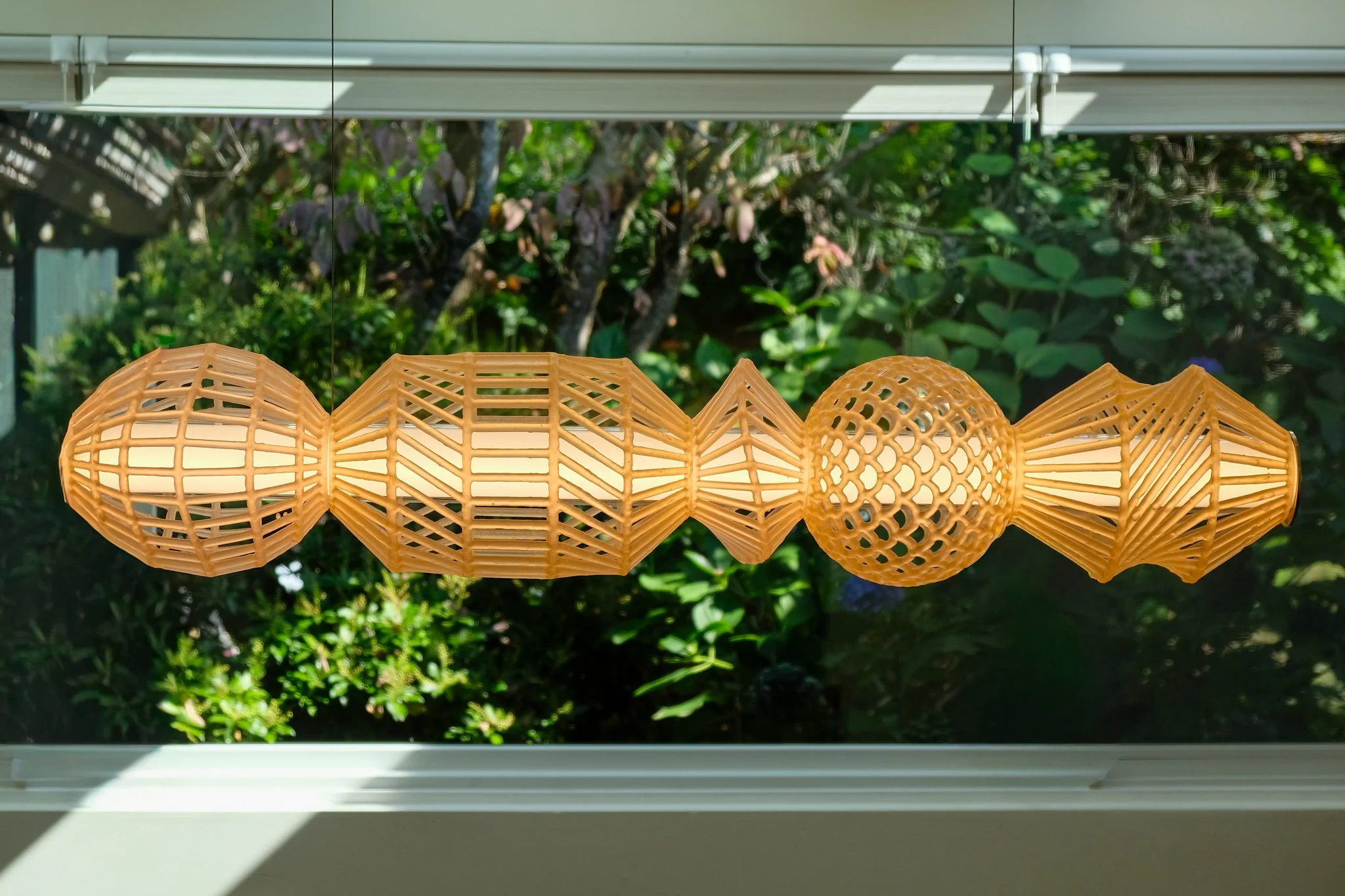 A decorative hanging lantern with intricate geometric patterns in a natural wood finish, hanging in front of a window with green foliage outside.