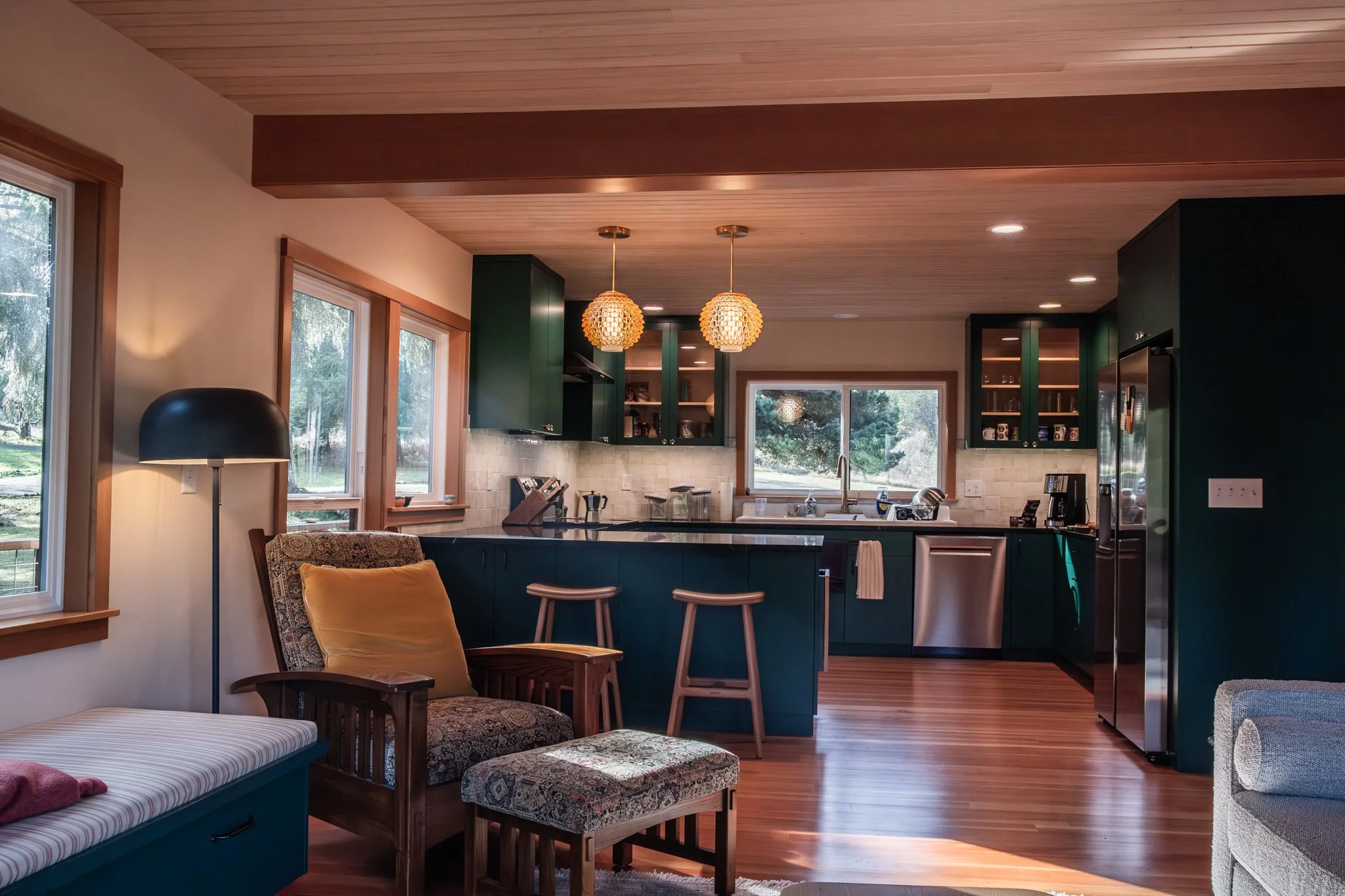 Cozy kitchen and living area with wood flooring, green cabinets, and large windows with a view of trees outside. There are two pendant lights, a chair with a yellow pillow, and a daily light lamp.