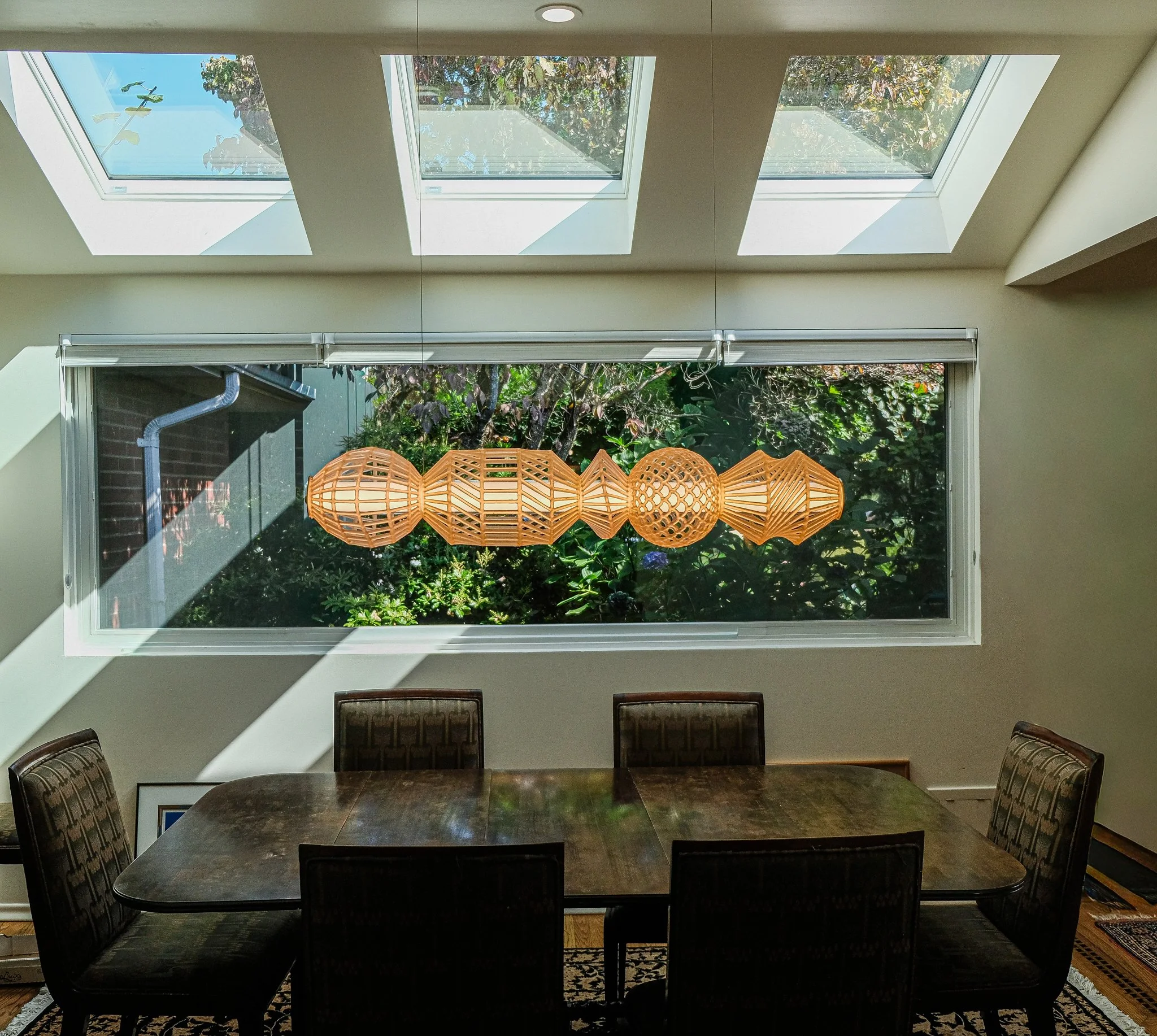 A dining room with a dark wooden table and six chairs. Three skylights in the ceiling and a large window behind the table. Hanging from the ceiling is a geometric wooden pendant light fixture.