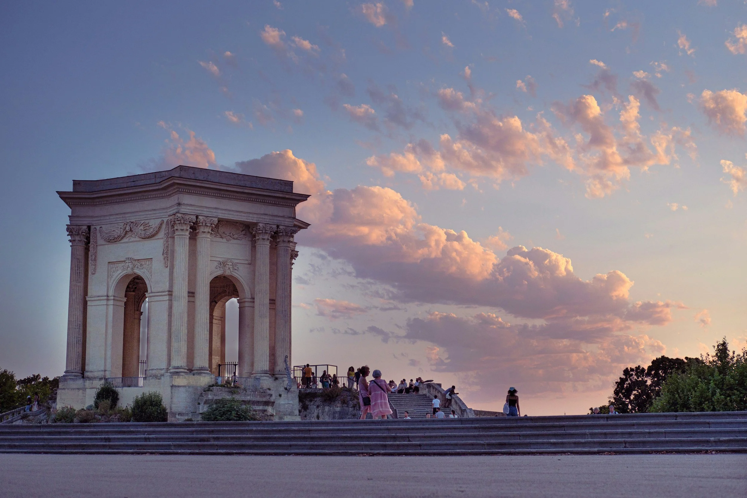 Vue d'une structure monumentale ornée de colonnes romaines, située en haut de marches avec des gens qui se baladent autour, sous un ciel au coucher du soleil avec des nuages