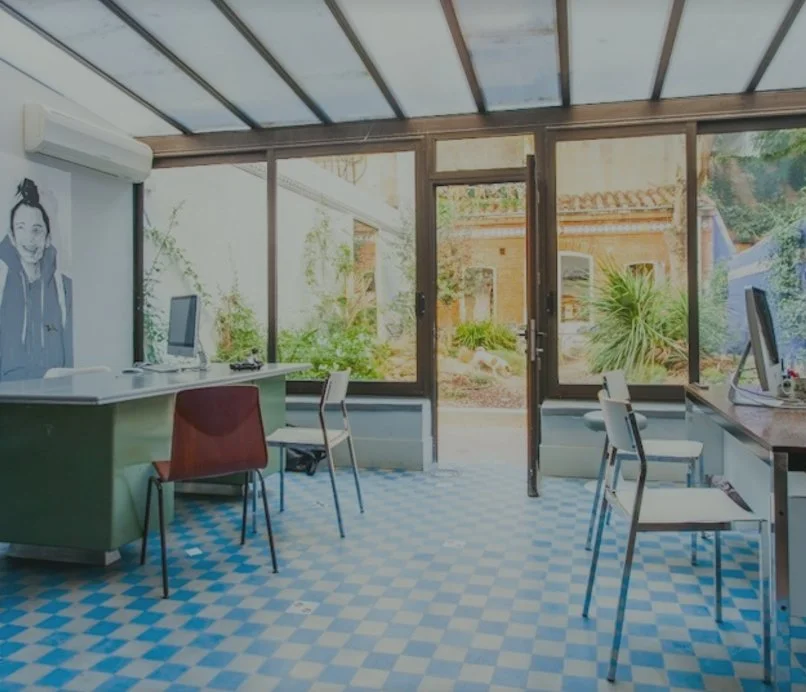 Une salle de bureau lumineuse avec des ordinateurs, des chaises, et de grandes fenêtres donnant sur un jardin extérieur.