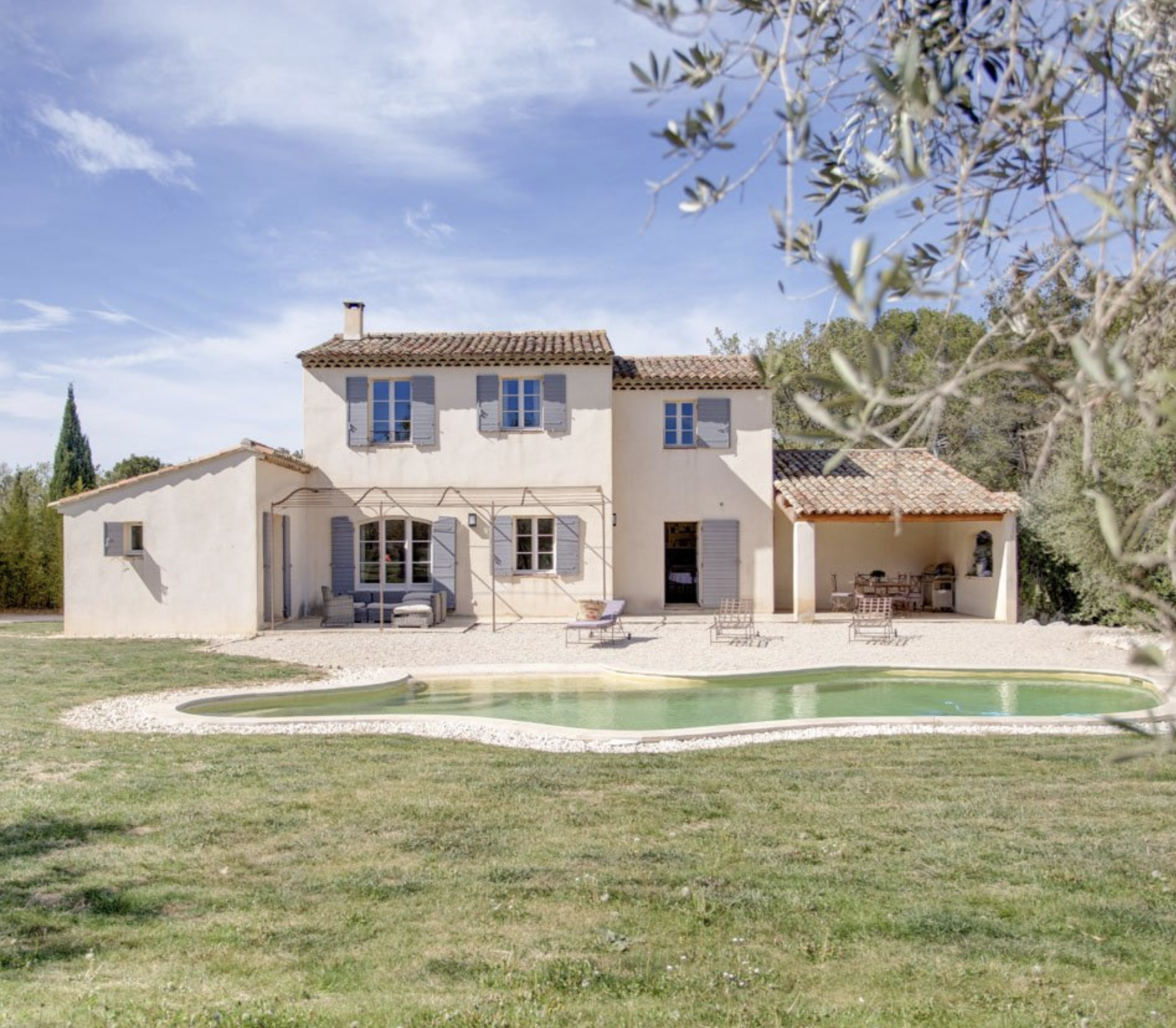 Maison de style méditerranéen avec piscine extérieure, terrasse avec mobilier et arbres environnants sous un ciel bleu clair.