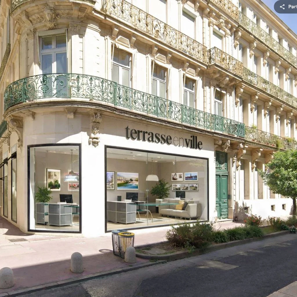 Vitrine d'une agence immobilière appelée 'terrasse en ville' située dans un bâtiment haussmannien avec balcons en fer forgé. La vitrine montre un intérieur moderne avec des bureaux, des plantes et des œuvres d'art.