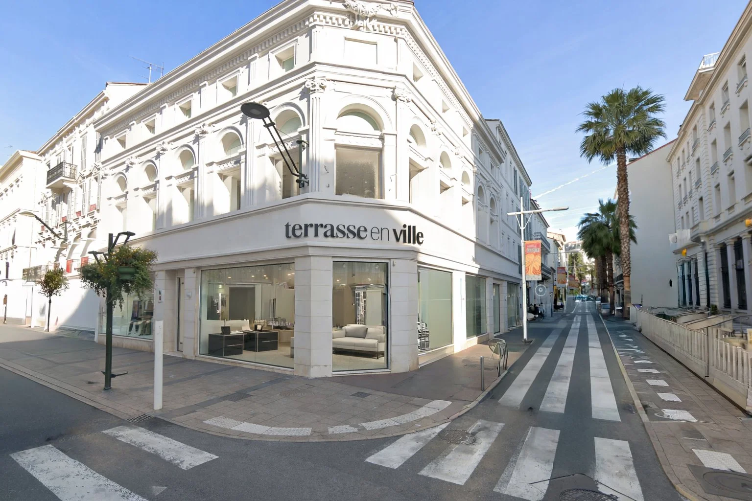 Vue d'une rue urbaine avec un bâtiment blanc à coins arrondis portant la signalisation 'terrasse en ville', des palmiers et un ciel clair.
