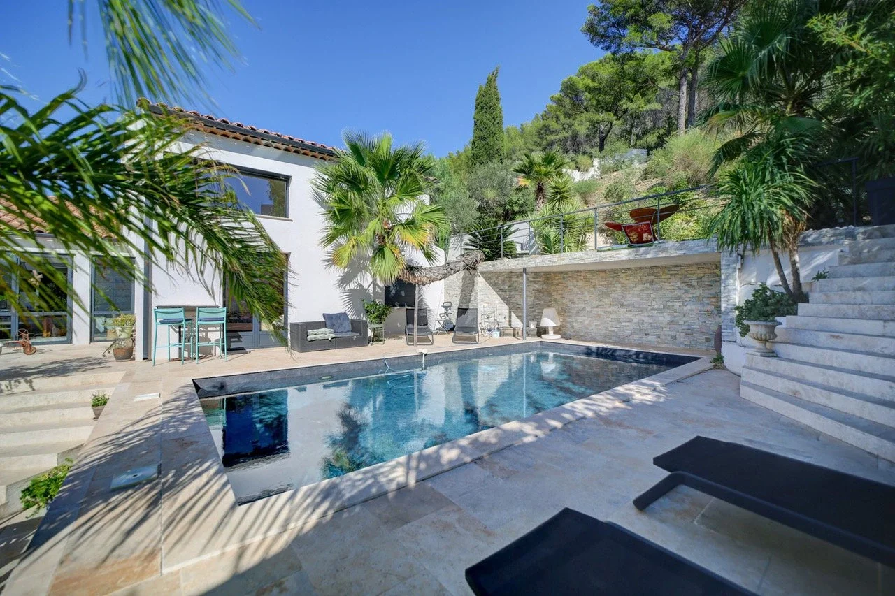 Piscine extérieure dans une cour avec plantes tropicales, chaises longues, et une maison moderne en arrière-plan.