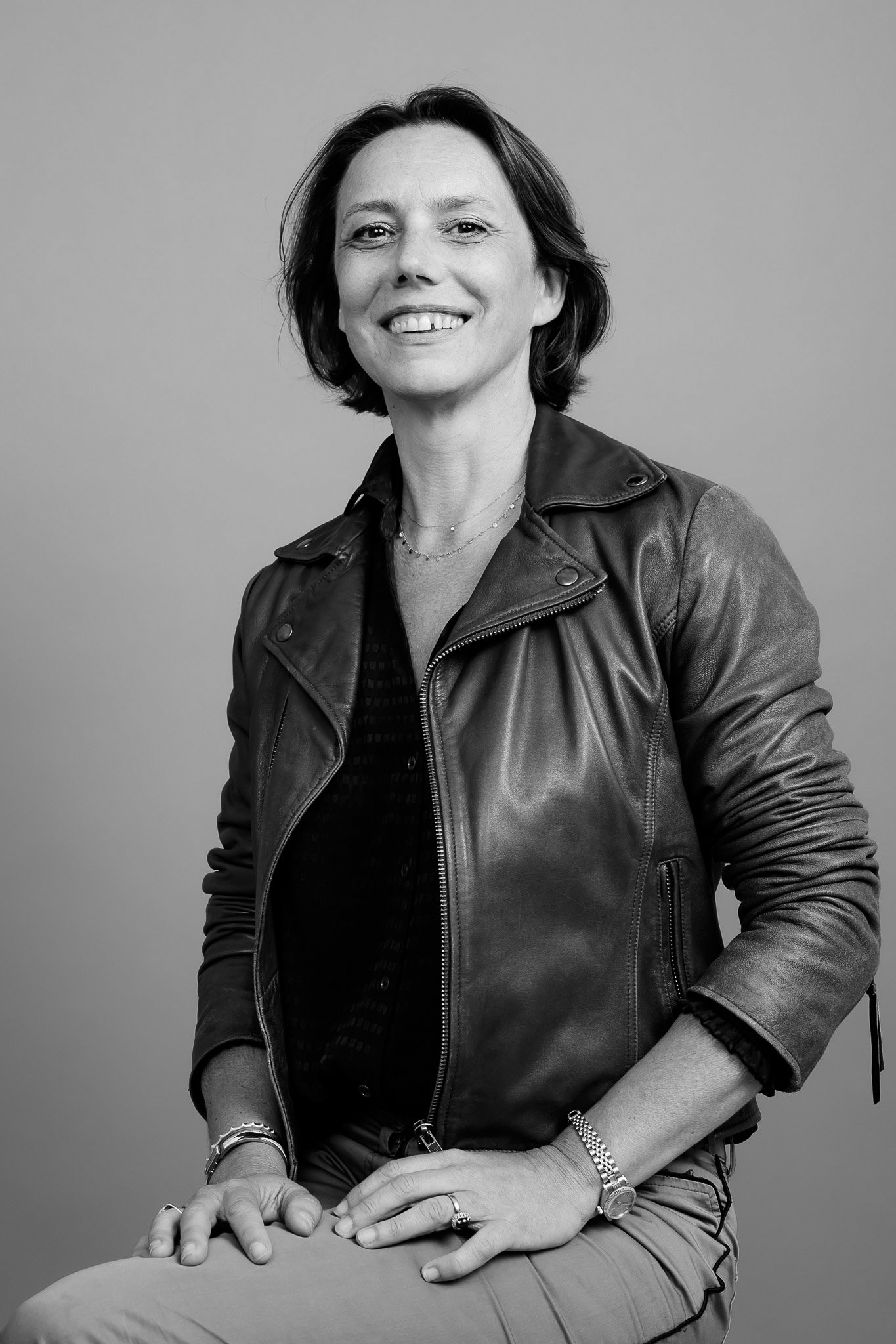 Portrait en noir et blanc d'une femme souriante, portant une veste en cuir, avec des bijoux et des bracelets, assise contre un fond simple.