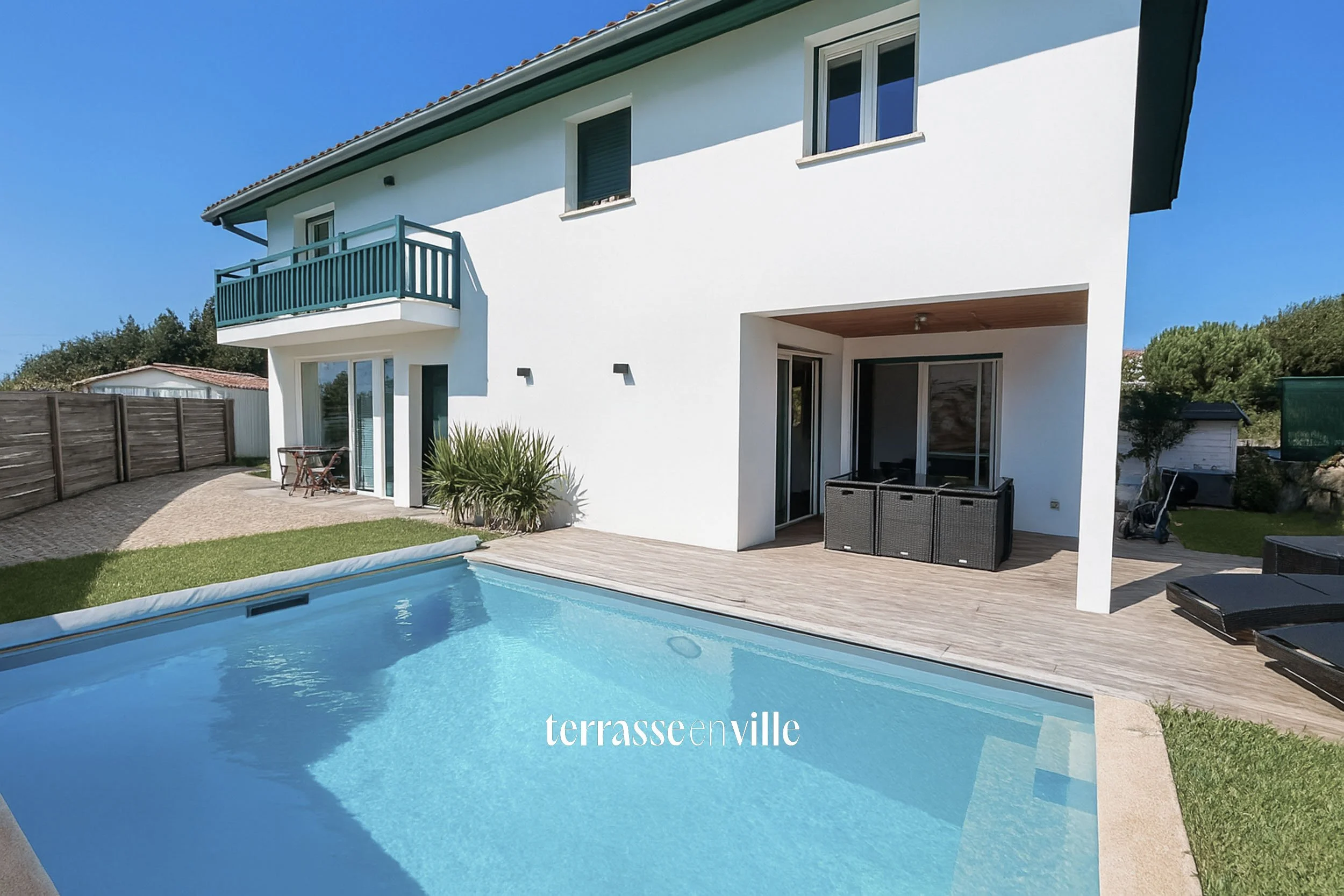 Vue d'une maison moderne avec piscine dans le jardin, terrasse en bois, balcon en haut avec une rambarde bleue, et mobilier extérieur