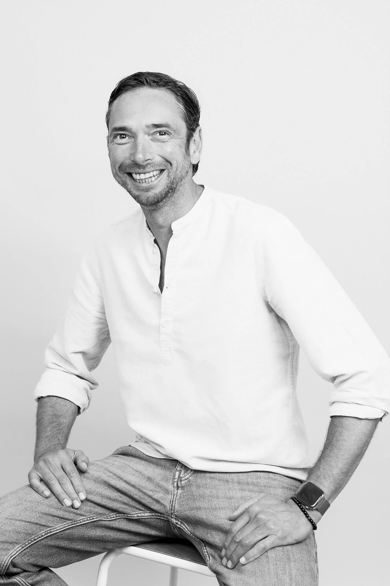 Un homme souriant, assis sur une chaise, portant une chemise blanche et un jean, dans un style noir et blanc.