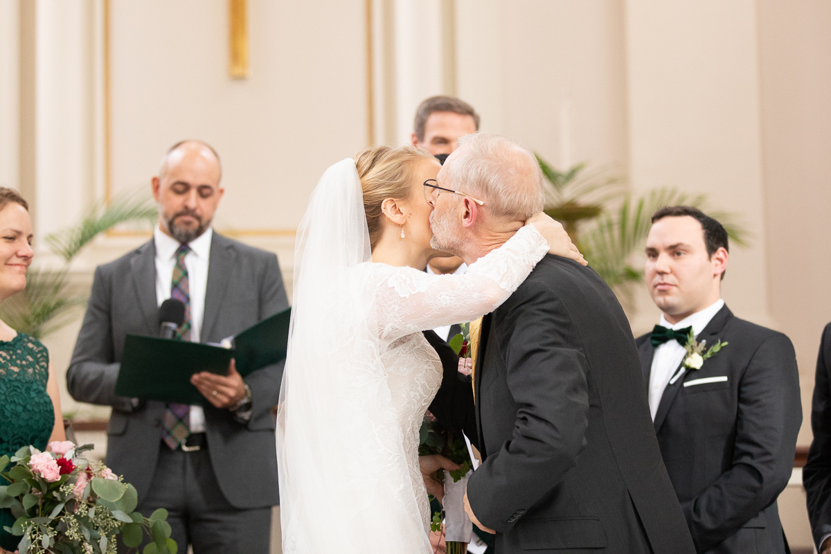 Bride’s father giving bride a kiss at wedding ceremony.