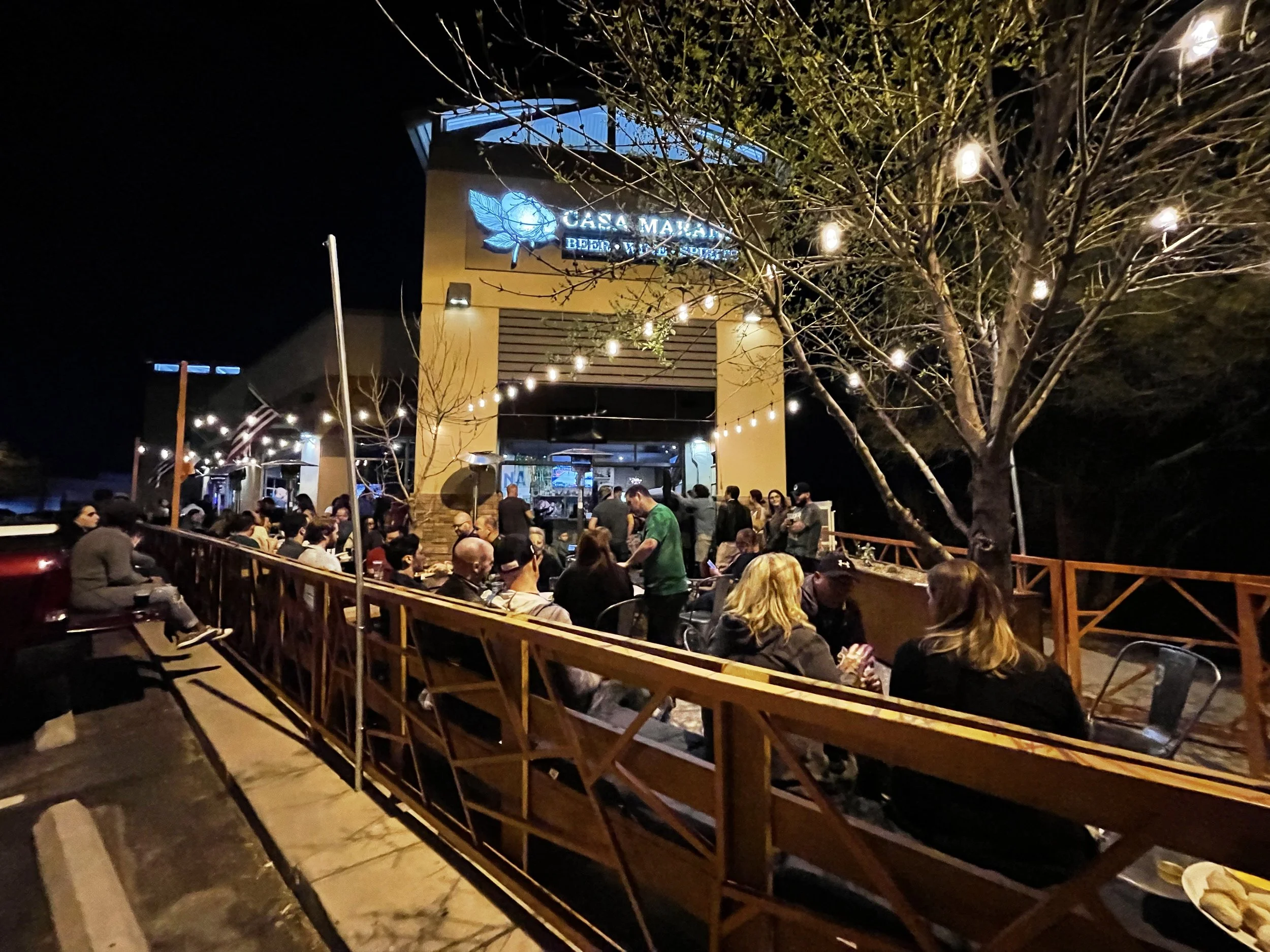 Night scene of a crowded outdoor patio or restaurant with string lights, trees, and a sign that reads 'Casa Mañana.'