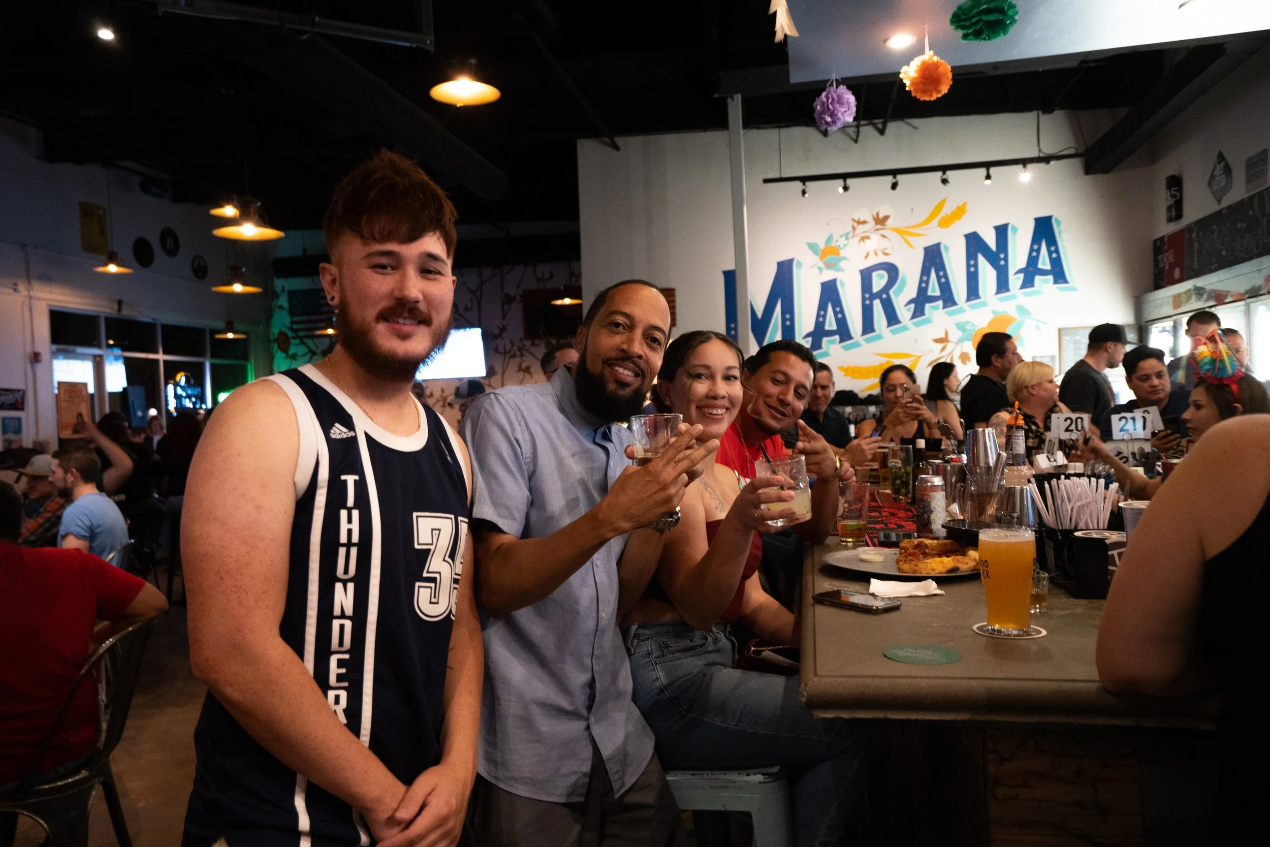 Group of five people sitting at a bar counter, smiling and holding drinks in a lively restaurant with colorful decorations and a large sign that reads 'Marana' in the background.