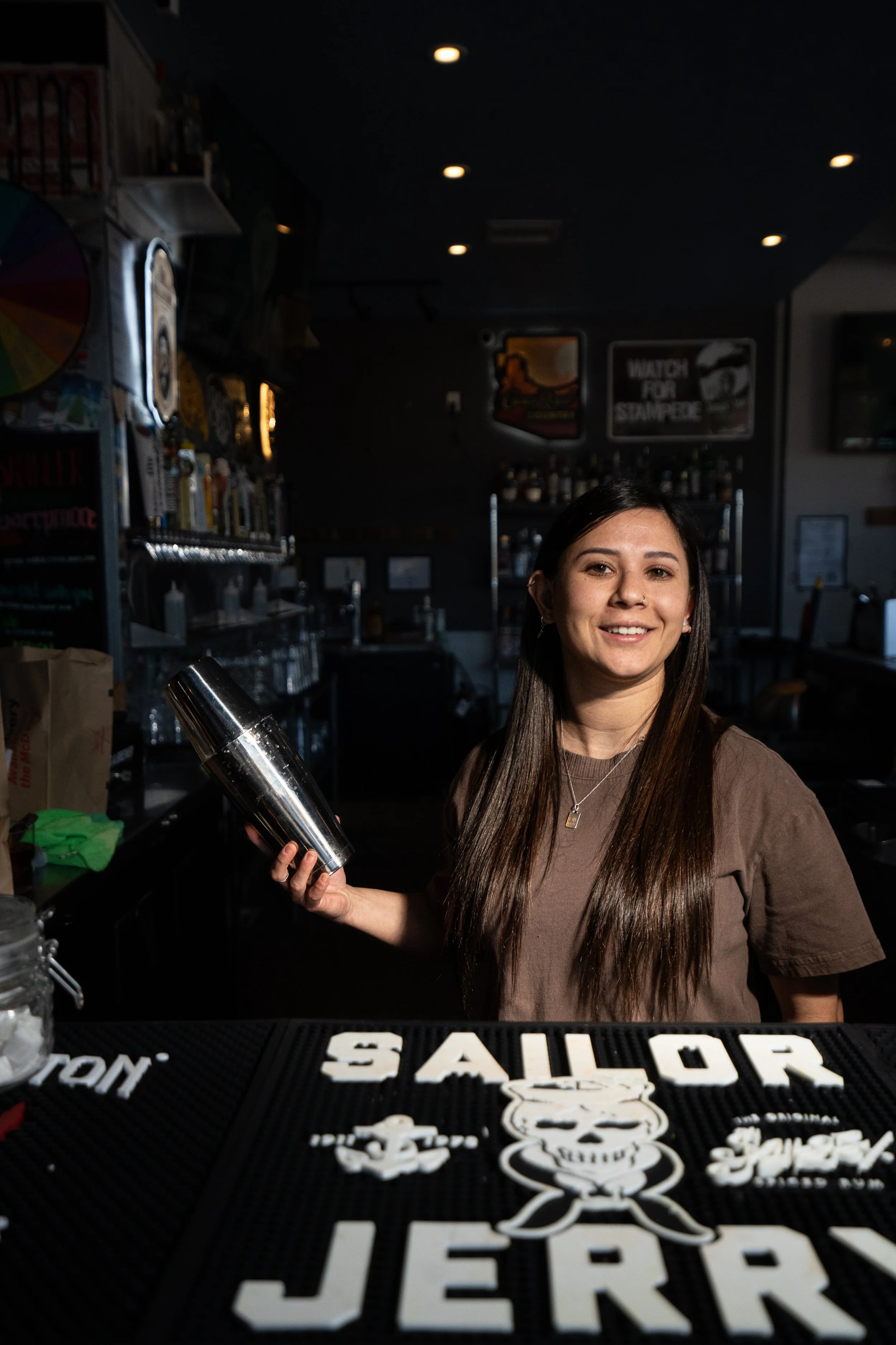A young woman with long brown hair smiling, holding a cocktail shaker behind a bar with a black bar mat that says 'JERK' and features a skull with sunglasses, in a dimly lit bar or pub setting.