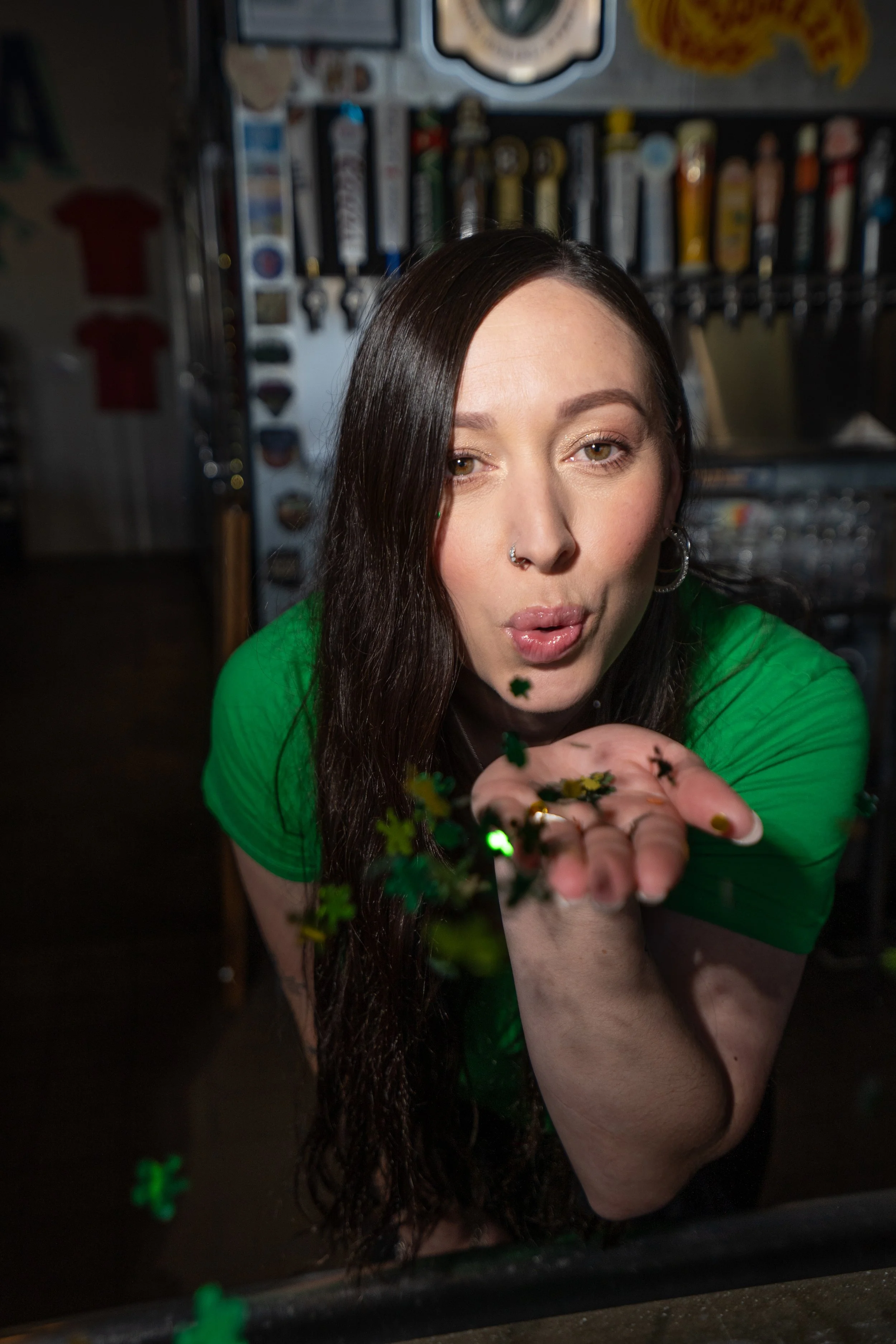 A woman with long dark hair, wearing a green shirt, is blowing green confetti towards the camera. She is in a bar or brewery, with beer taps in the background.