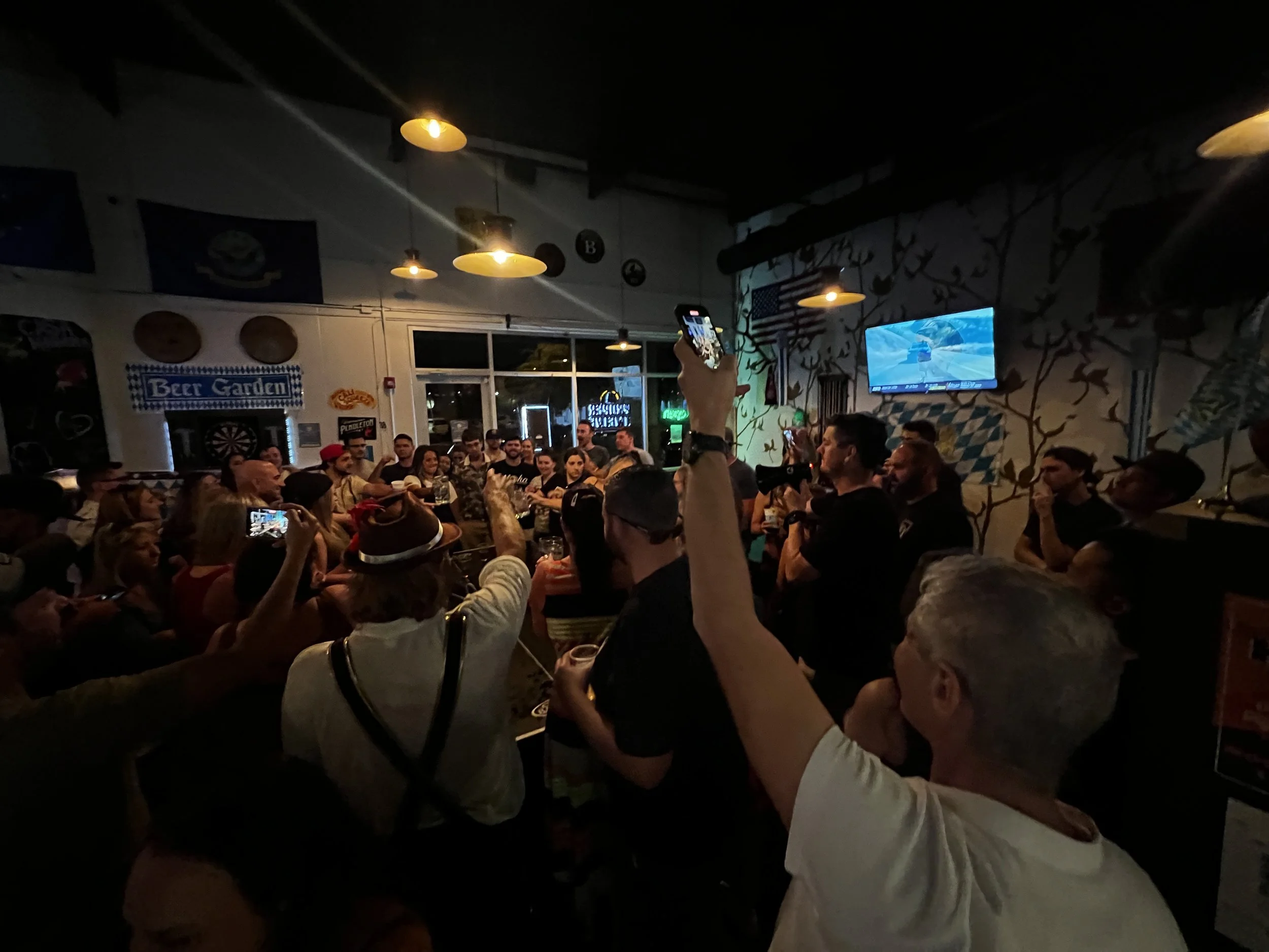 A crowded bar with people watching a television and taking photos, with warm lighting and wall decorations.