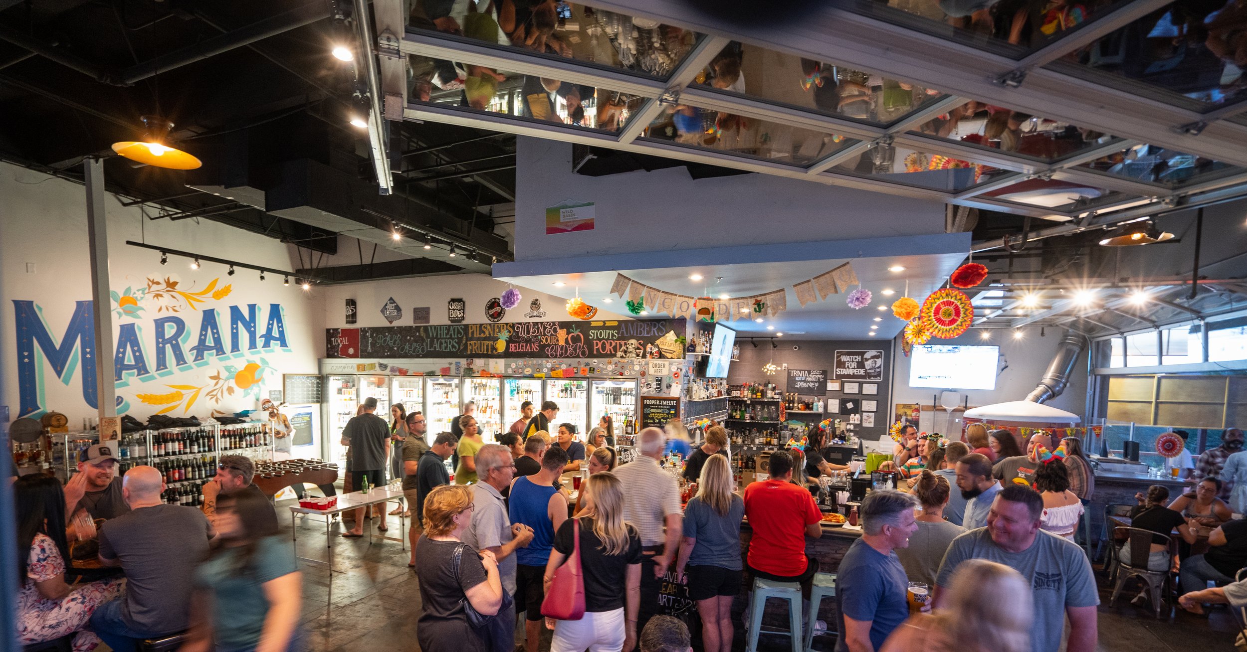 A crowded indoor bar or brewery with people socializing, colorful decorations like paper flowers, and a mural on the wall that says 'MARANA'. The ceiling has mirrors reflecting the crowd and lights, and there are menu boards and a TV screen visible i