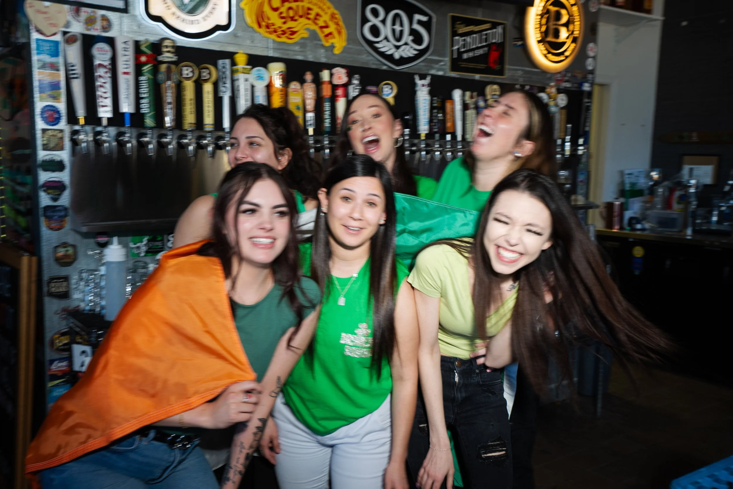 A group of young women laughing and smiling, standing close together in front of a bar with beer taps and beer-related signs.