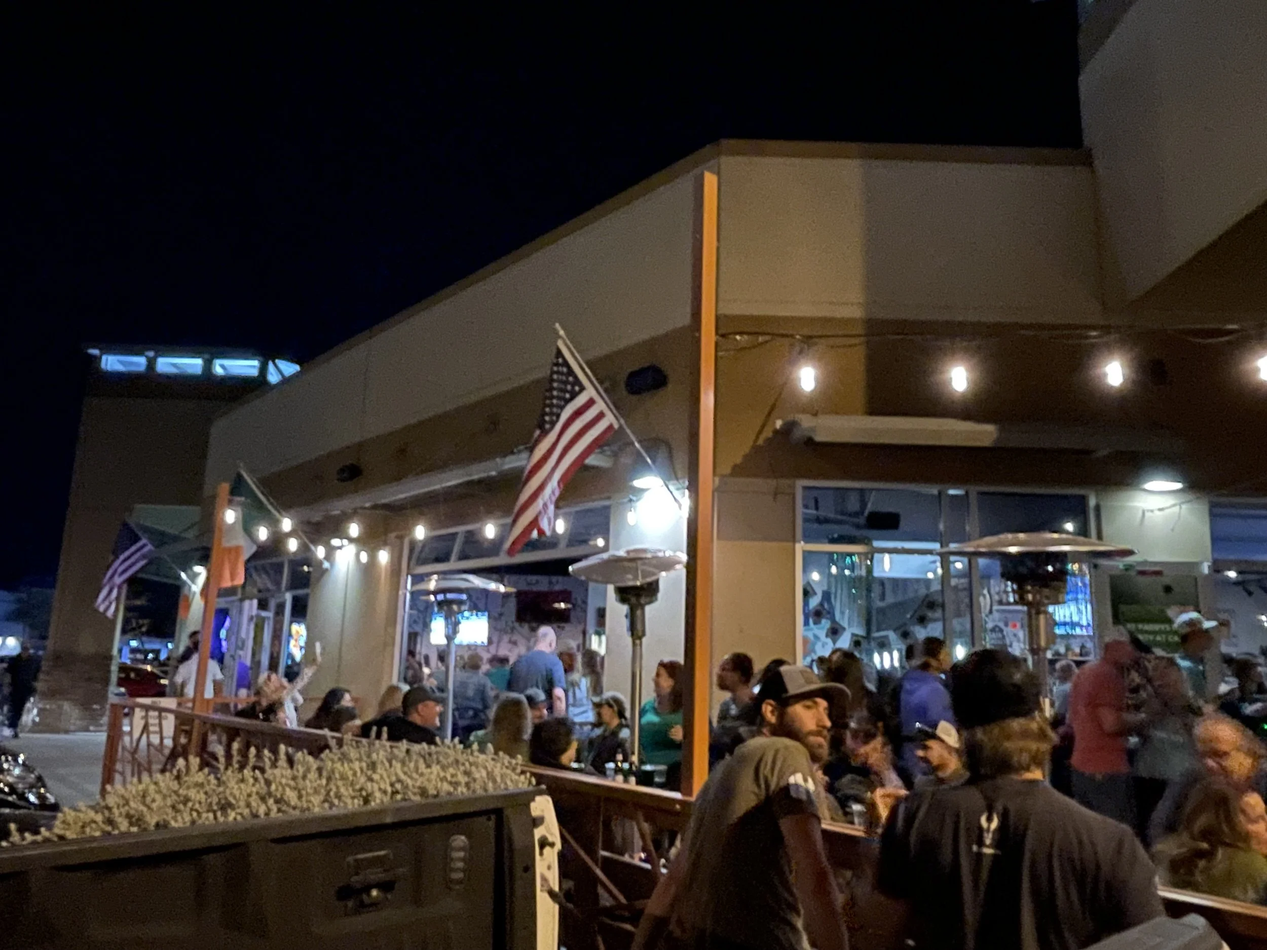 Nighttime scene of an outdoor restaurant or bar with people seated at tables, American flags, string lights, and a building with a modern design and large windows.