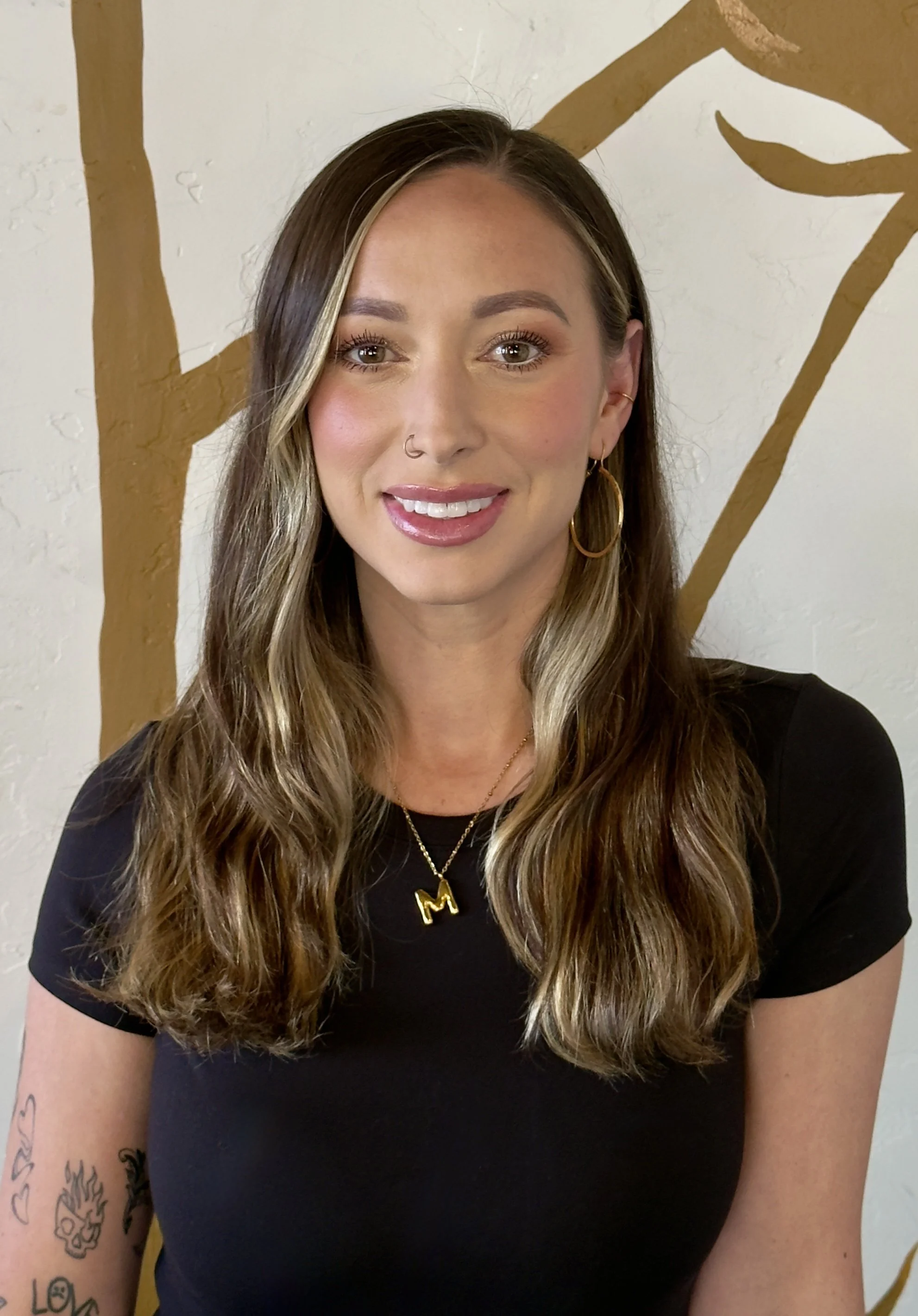 A young woman with long, wavy brown hair with blonde highlights, smiling, wearing a black t-shirt, gold hoop earrings, a gold necklace with an 'M' pendant, and a nose ring. She has tattoos on her left arm, and a painted tree branch with leaves is visible on the wall behind her.
