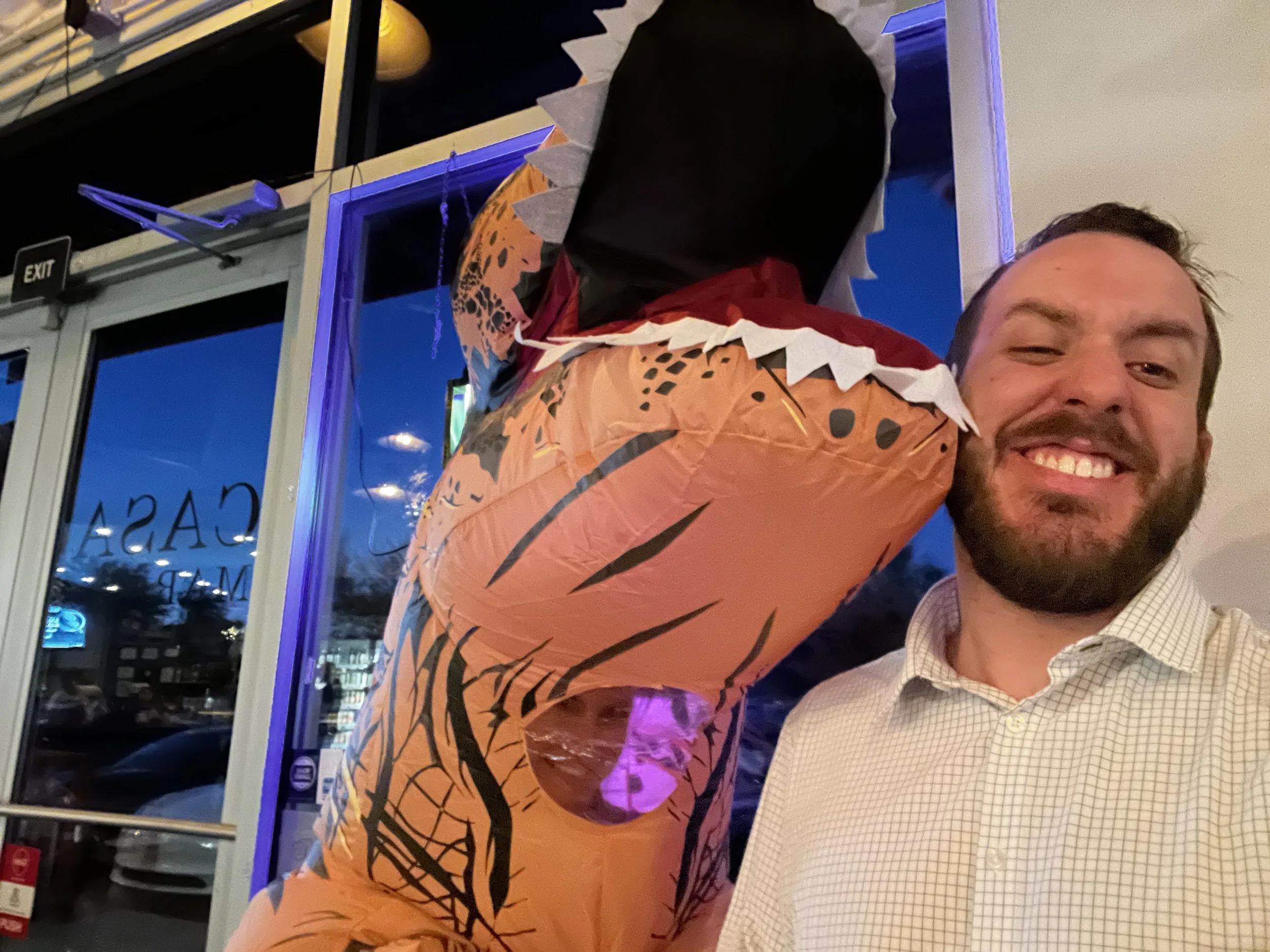 A man smiling while holding an inflatable dinosaur costume with orange and black striped pattern and spikes on its back, standing indoors near a window and a door.