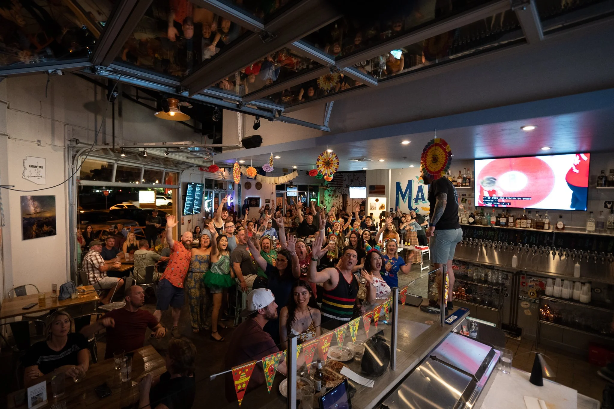 A lively bar or restaurant filled with many people celebrating, with colorful papel picado decorations hanging from the ceiling, and a person standing on a bar counter. The crowd looks cheerful and is raising their hands, and the setting is vibrant a