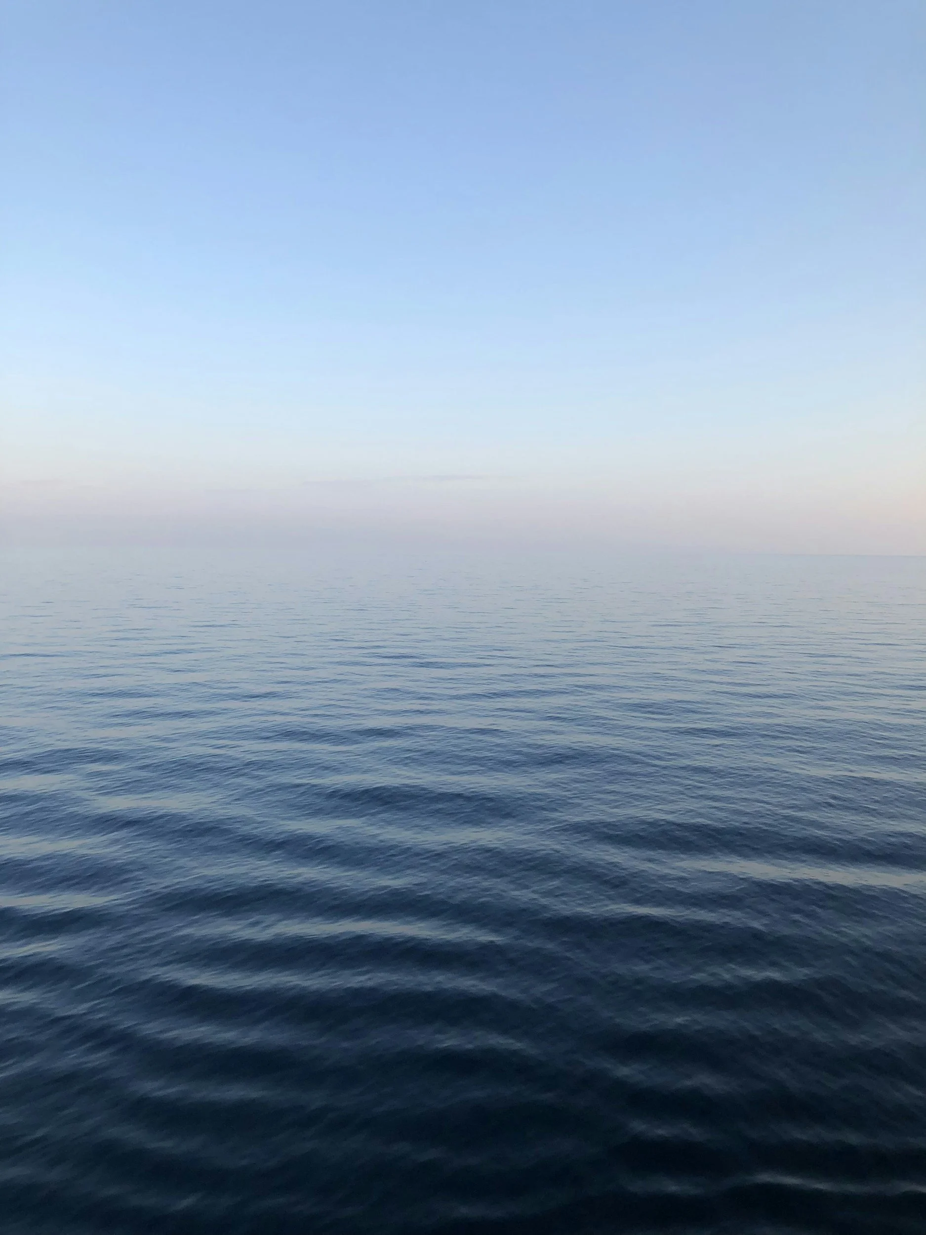 Calm ocean with gentle waves, clear blue sky, and a subtle gradient from light to dark blue near the horizon.
