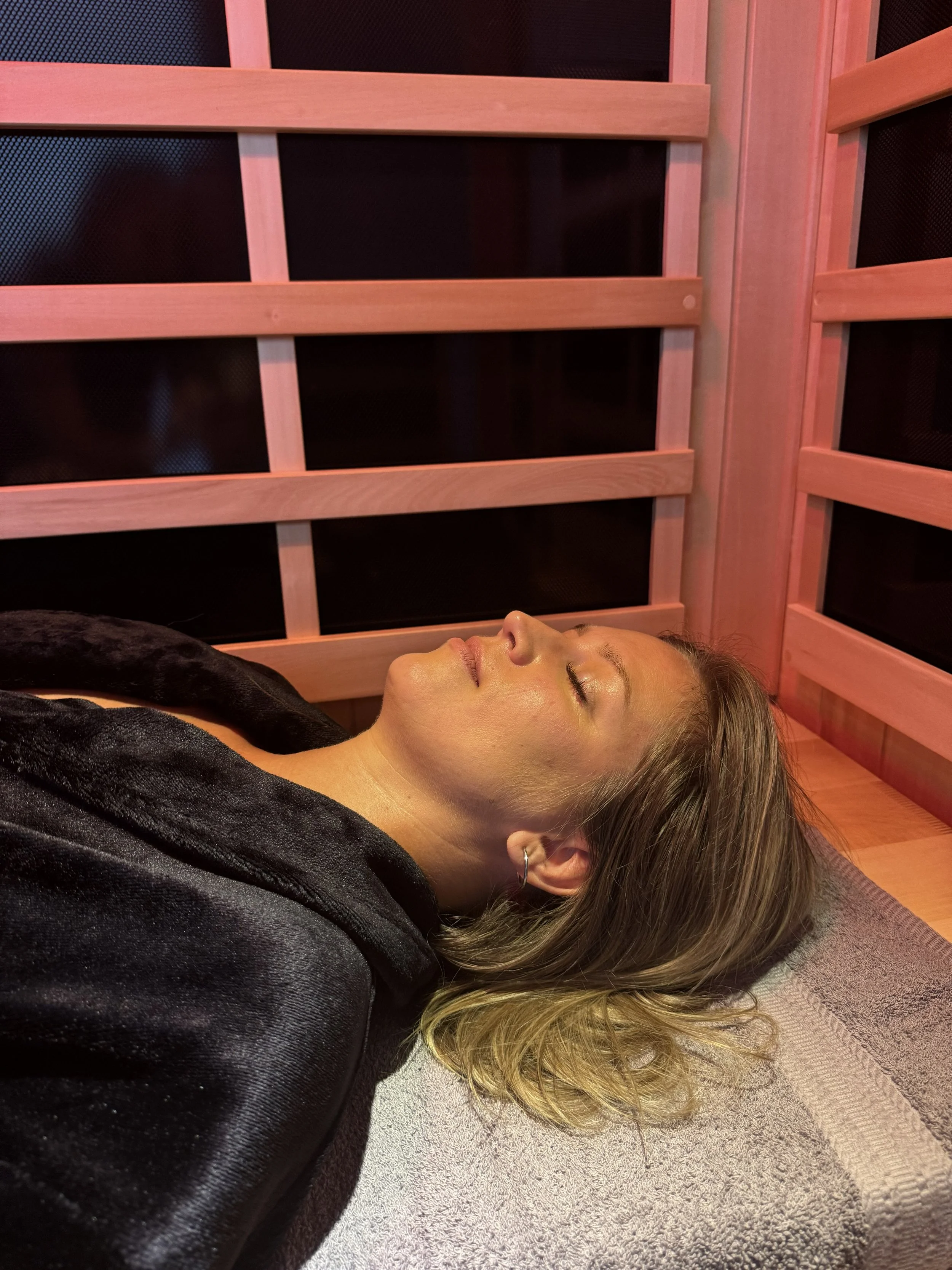 A woman lying down with her eyes closed inside a wooden sauna, wrapped in a black towel.