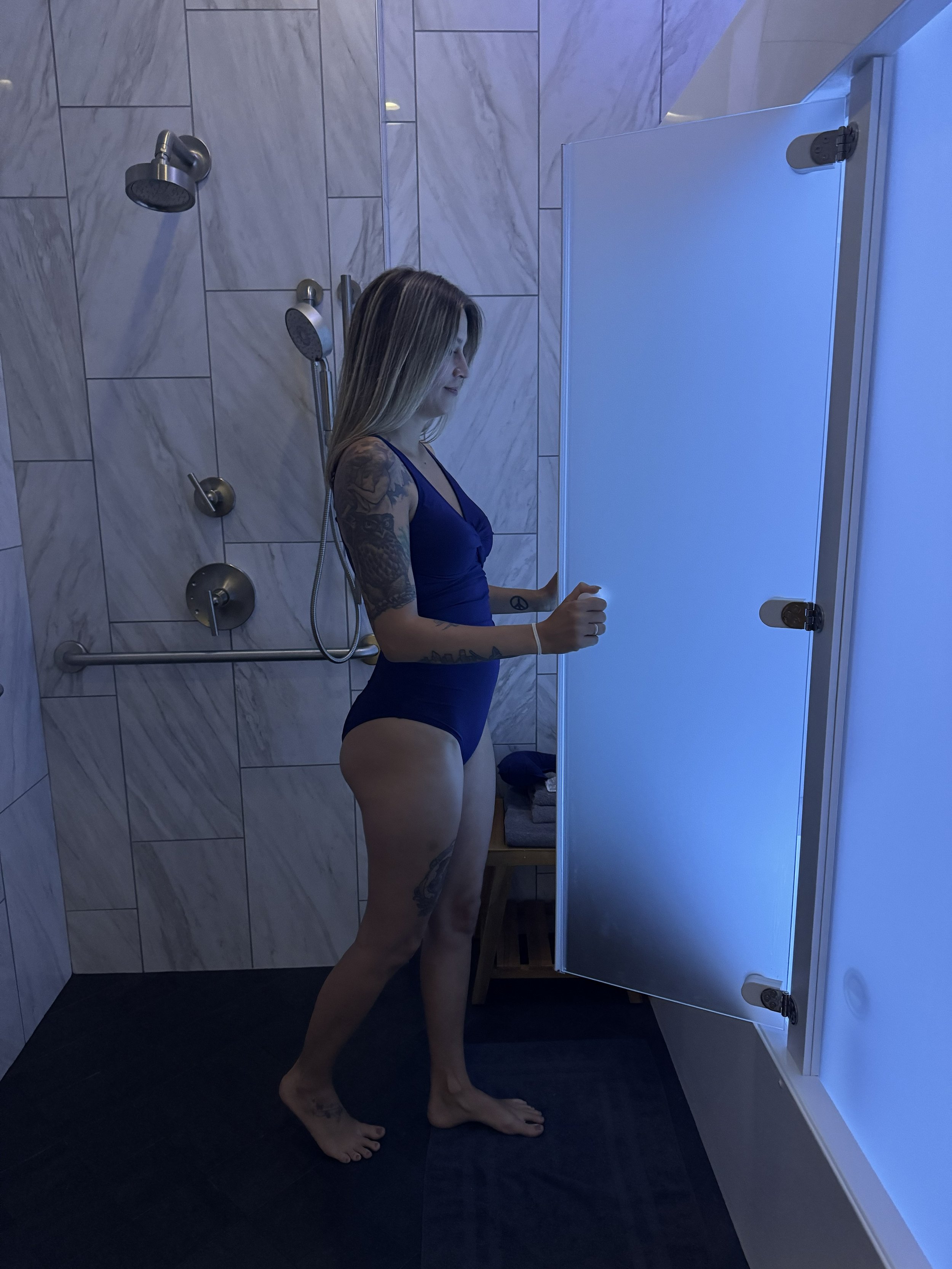 A woman with tattoos in a blue swimsuit standing in a shower area with tiled walls, holding door to a float tank.