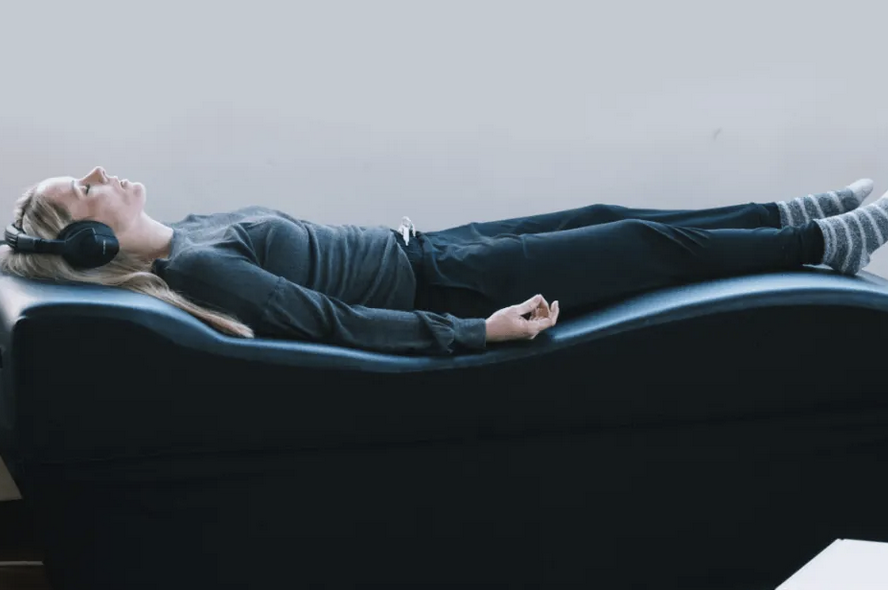 A woman lying on her back on a black medical exam table, wearing headphones, a gray long-sleeve shirt, dark pants, and striped socks, appearing to relax or meditate on the vibroacoustic soundbed.