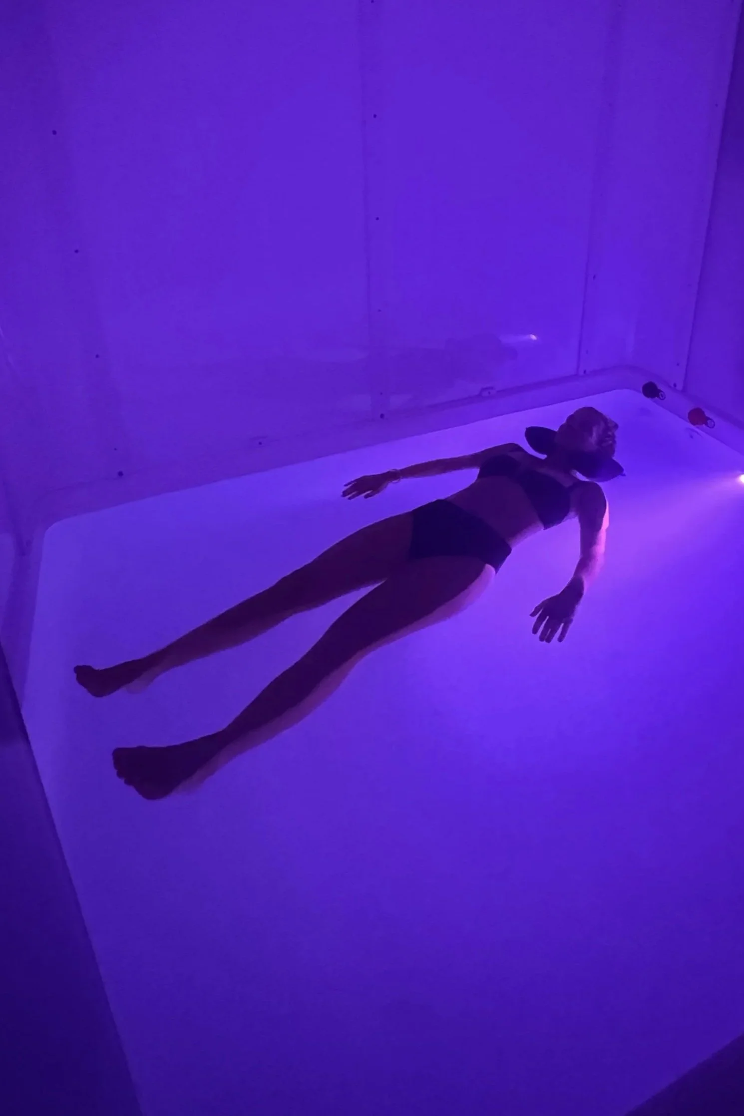 A person lying on a surface on the float tank water illuminated with purple and blue lighting in a dark room.