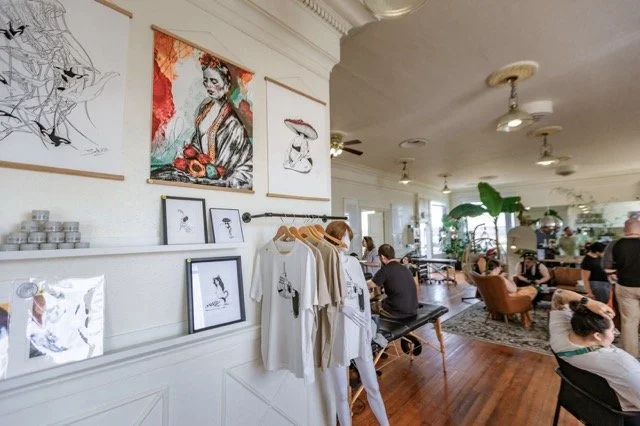Interior of a café or art space with artwork on the walls, a clothing rack with T-shirts, and people sitting and socializing.