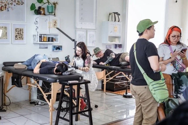 People lying on massage tables receiving massages while others stand and look at their phones in a bright, modern room with white walls and framed artwork.