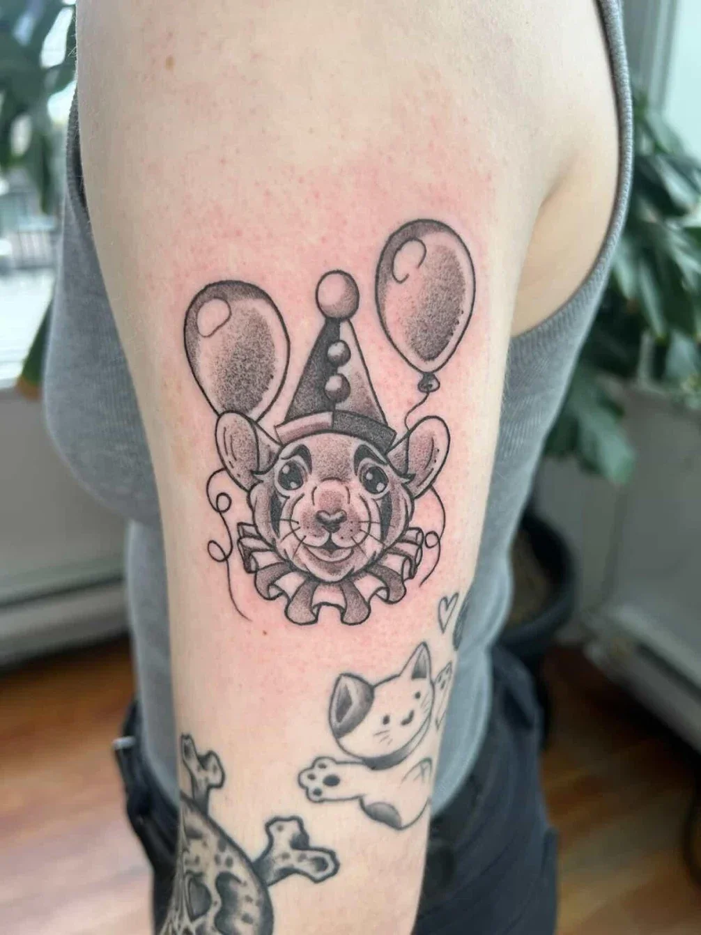 Tattoo of a cute animal with a party hat and balloons, designed as a clown or festive character, on a person's arm.