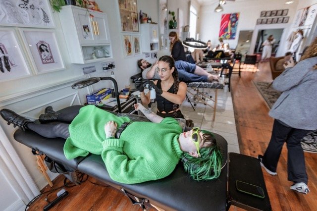Multiple people receiving tattoos in an art studio. A person with green hair and sunglasses is lying on a tattoo bed, while others are sitting or standing nearby.