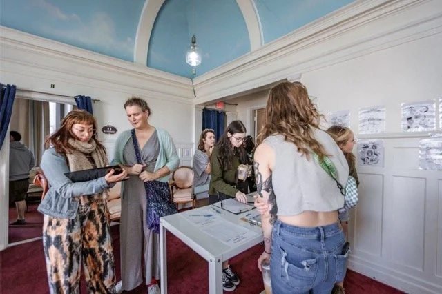 Group of women and girls at an indoor art exhibit or event, with artwork displayed on the walls and a white table in the center.