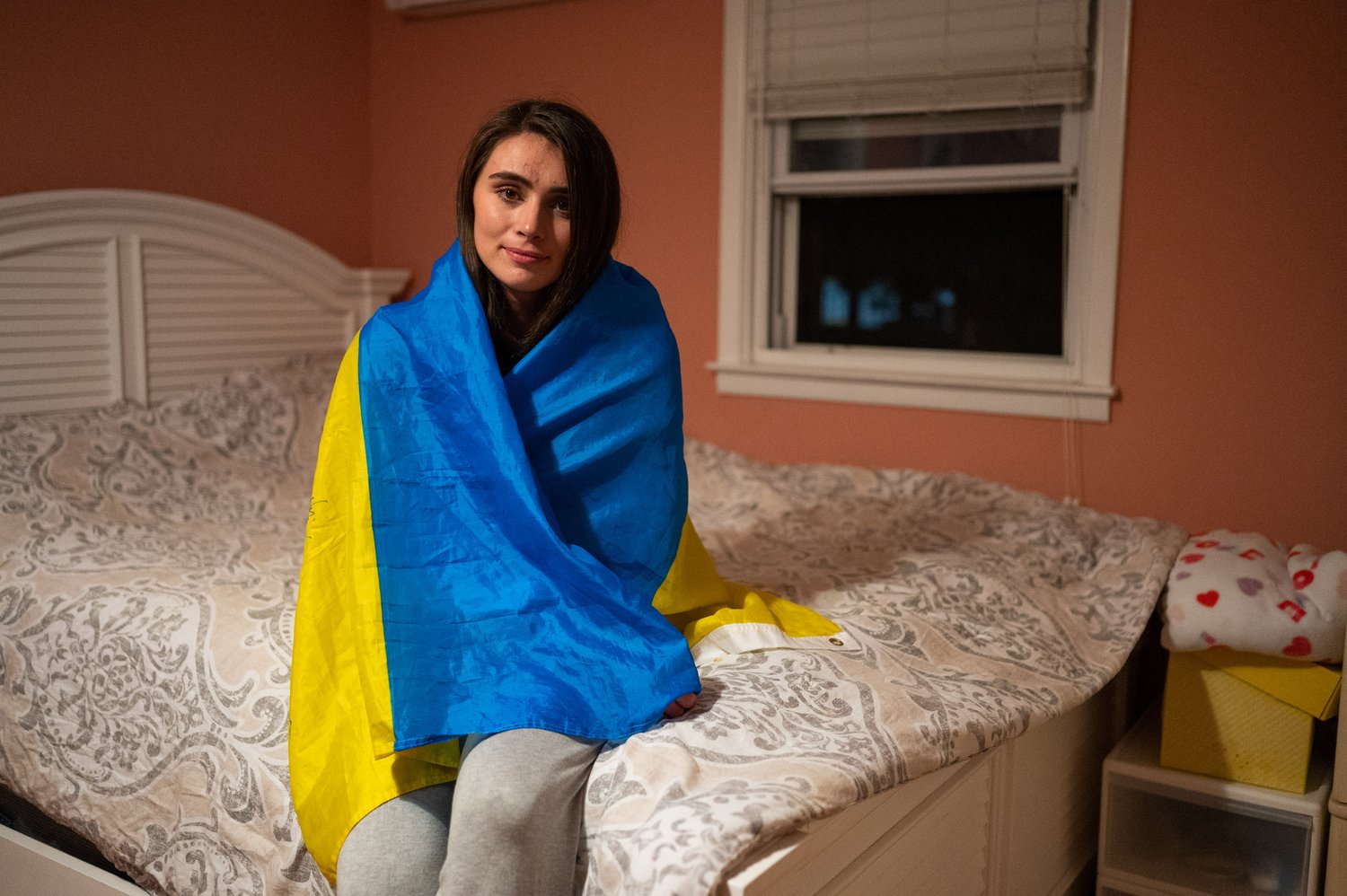   Yuliia Bihun drapes herself with the Ukrainian flag while sitting on her bed.  