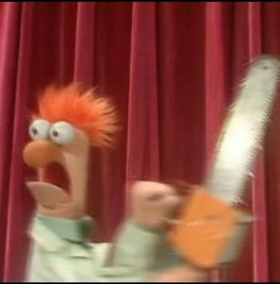 A puppet with orange hair, wide eyes, and an open mouth, holding an orange object in front of a red curtain.