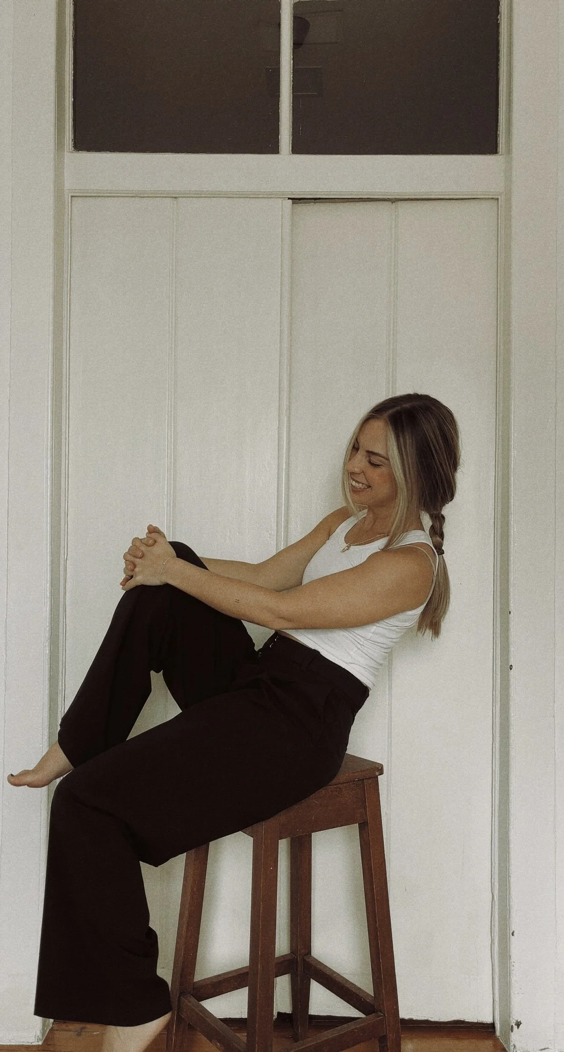 A woman sitting on a wooden stool with her leg up, smiling and holding her knee, against a white paneled wall with a window above.
