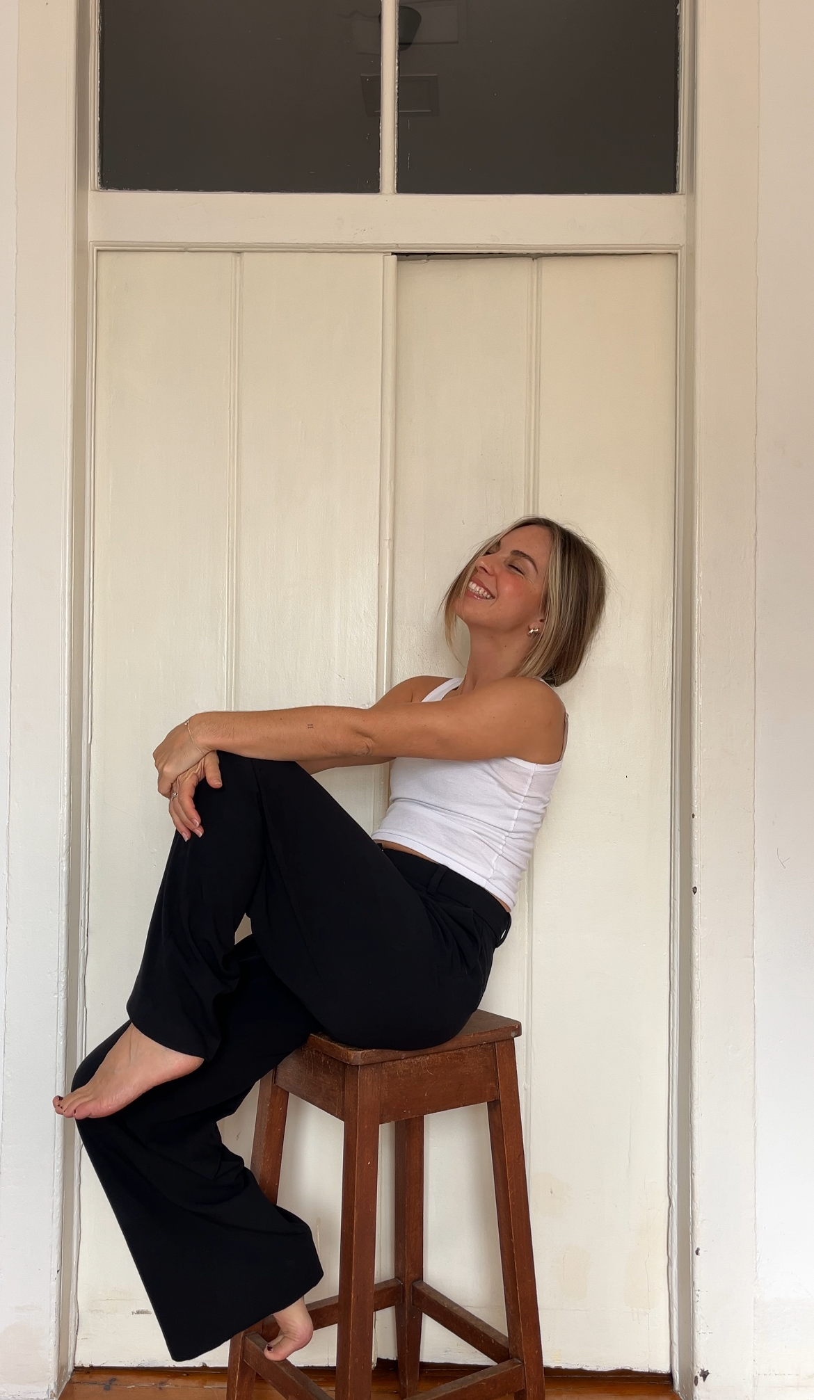 A woman sitting on a wooden stool against a white wall with a small window above. She is smiling with eyes closed, wearing a white tank top and black pants.
