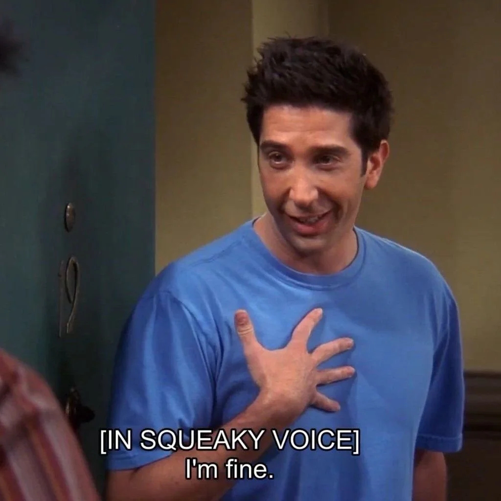 A man with dark, spiky hair and wearing a blue t-shirt is touching his chest with his right hand and smiling. There are subtitles that say '[IN SQUEAKY VOICE] I'm fine.'