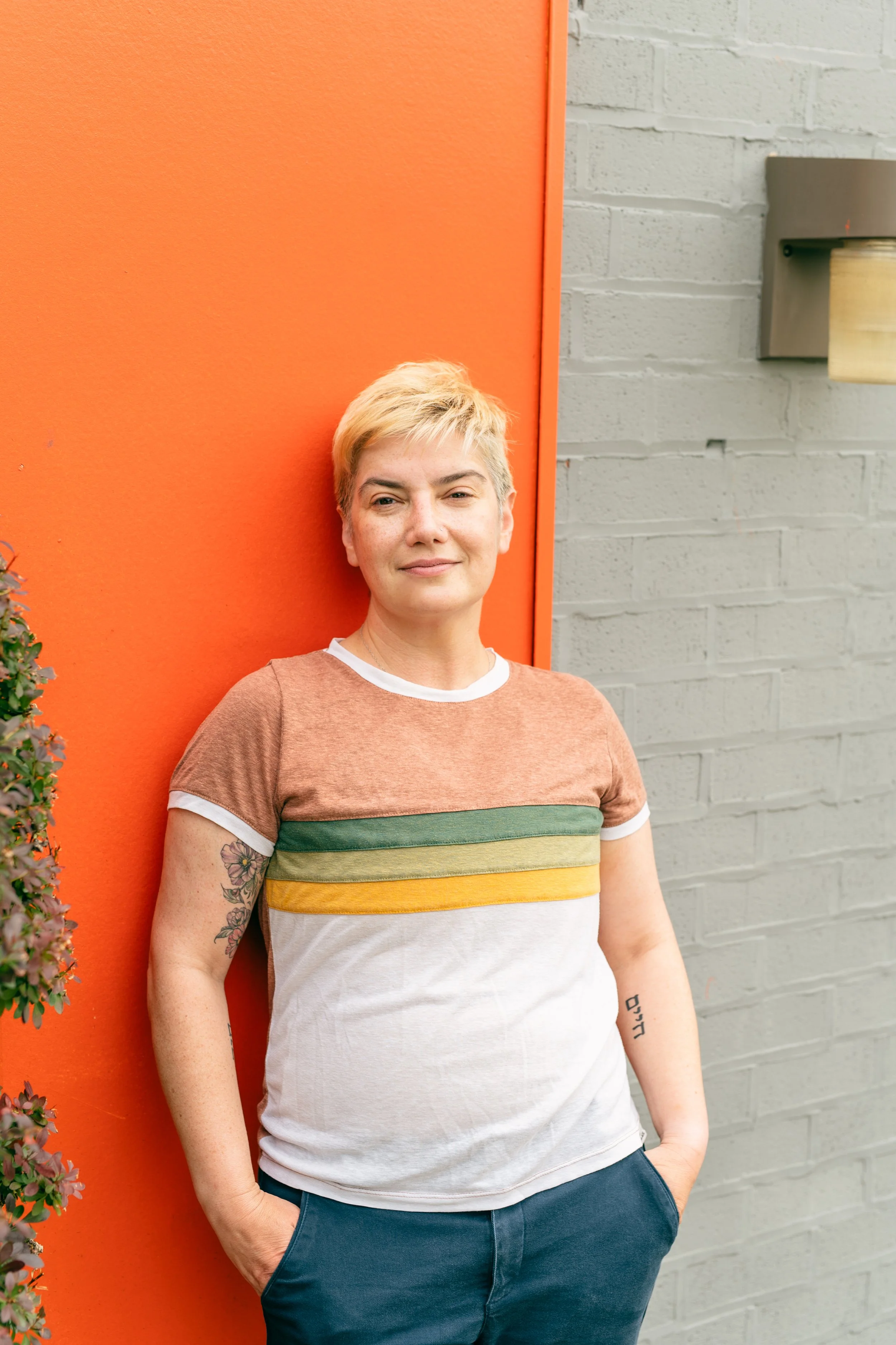 A white nonbinary person with a boyish blond haircut leans, with hands in pockets, against an orange wall. They wear a white, yellow, green, and mauve t-shirt and dark blue chinos.