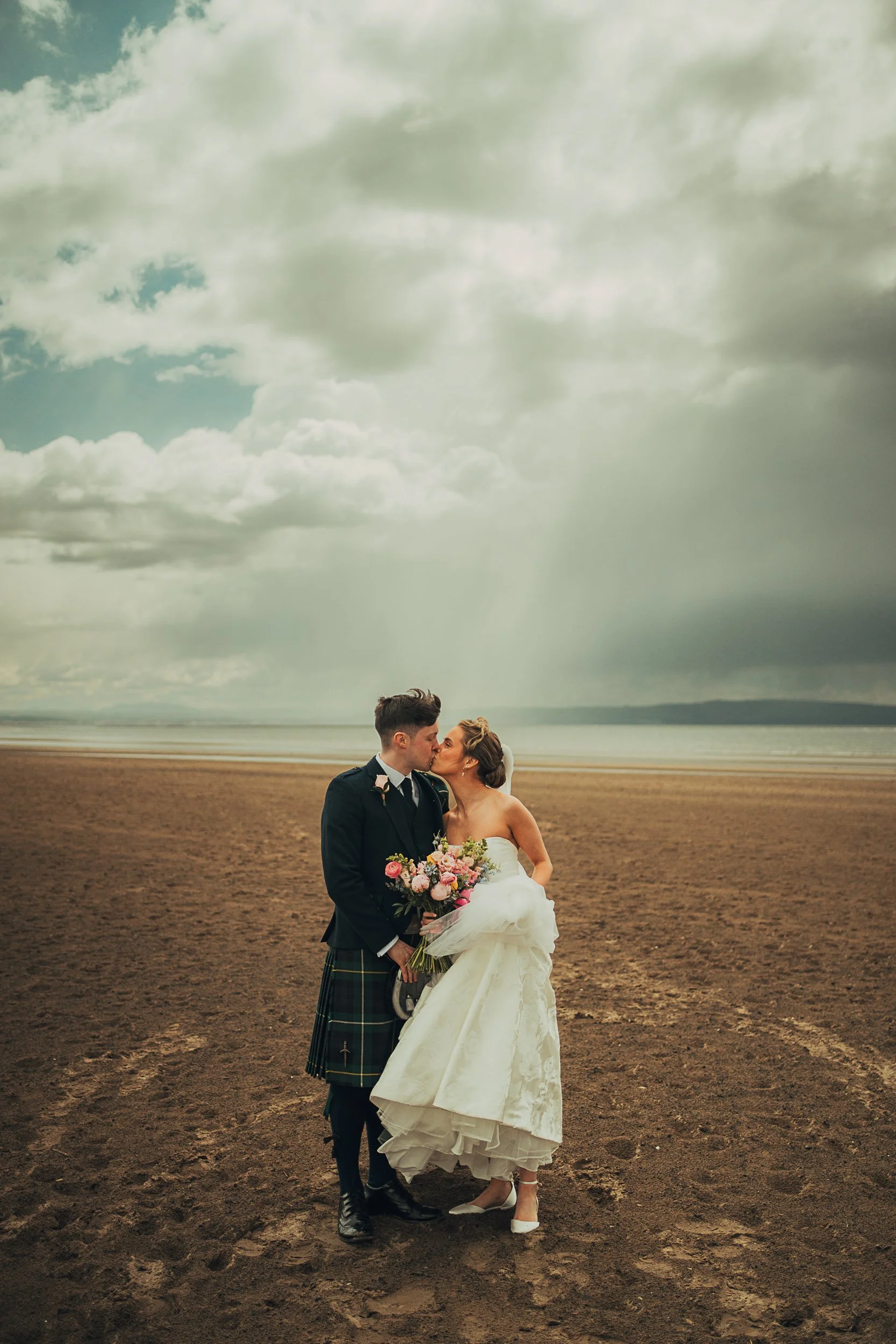 Bride and groom kissing on Troon beach beneath a dramatic Scottish sky after their wedding ceremony.