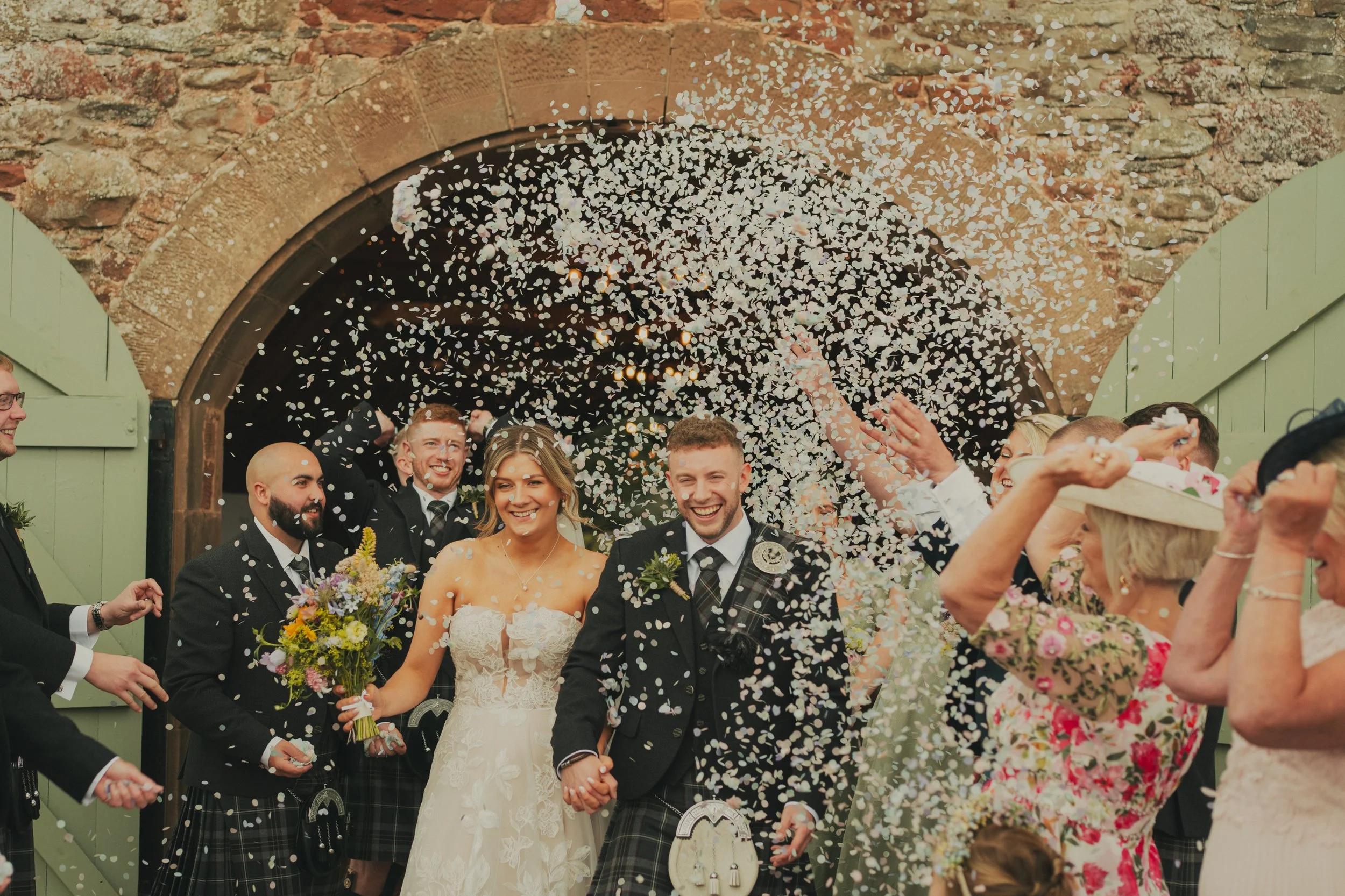 Bryony and Ryan walking through confetti beneath the courtyard archway at Wedderlie House