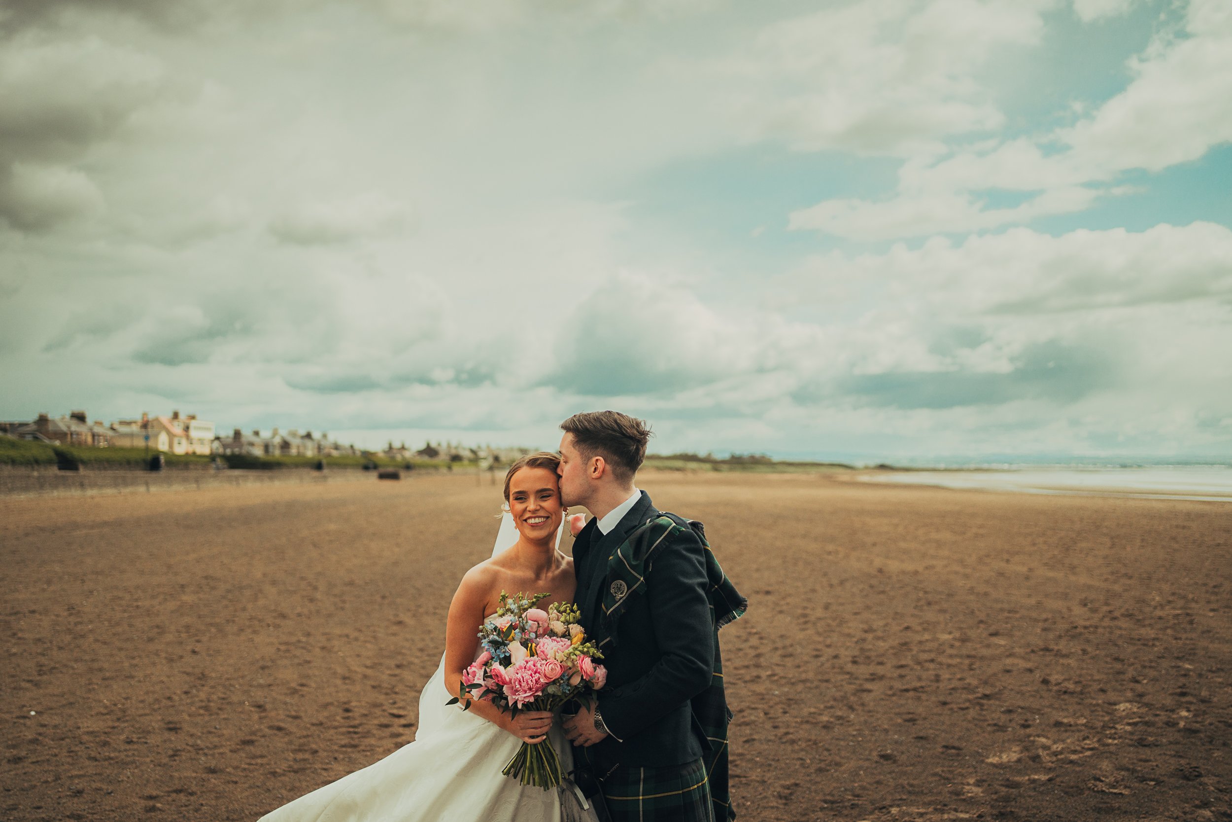 Groom kissing bride on the forehead during a relaxed couple shoot on Troon beach after their Lochgreen House Hotel wedding in Scotland.