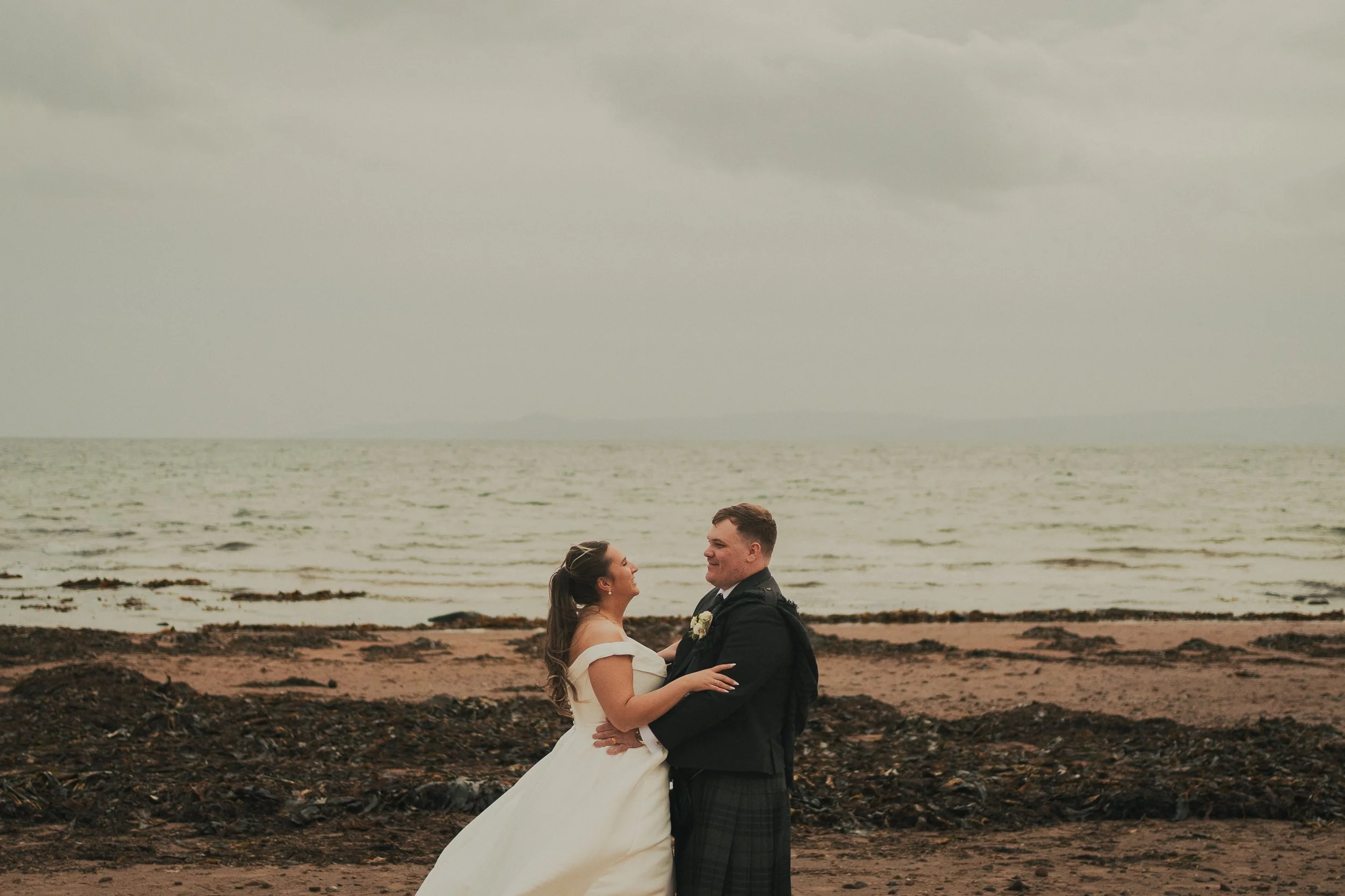 Gill and Callum looking at each other arm in arm on the beach at their Seamill Hydro wedding in Ayrshire